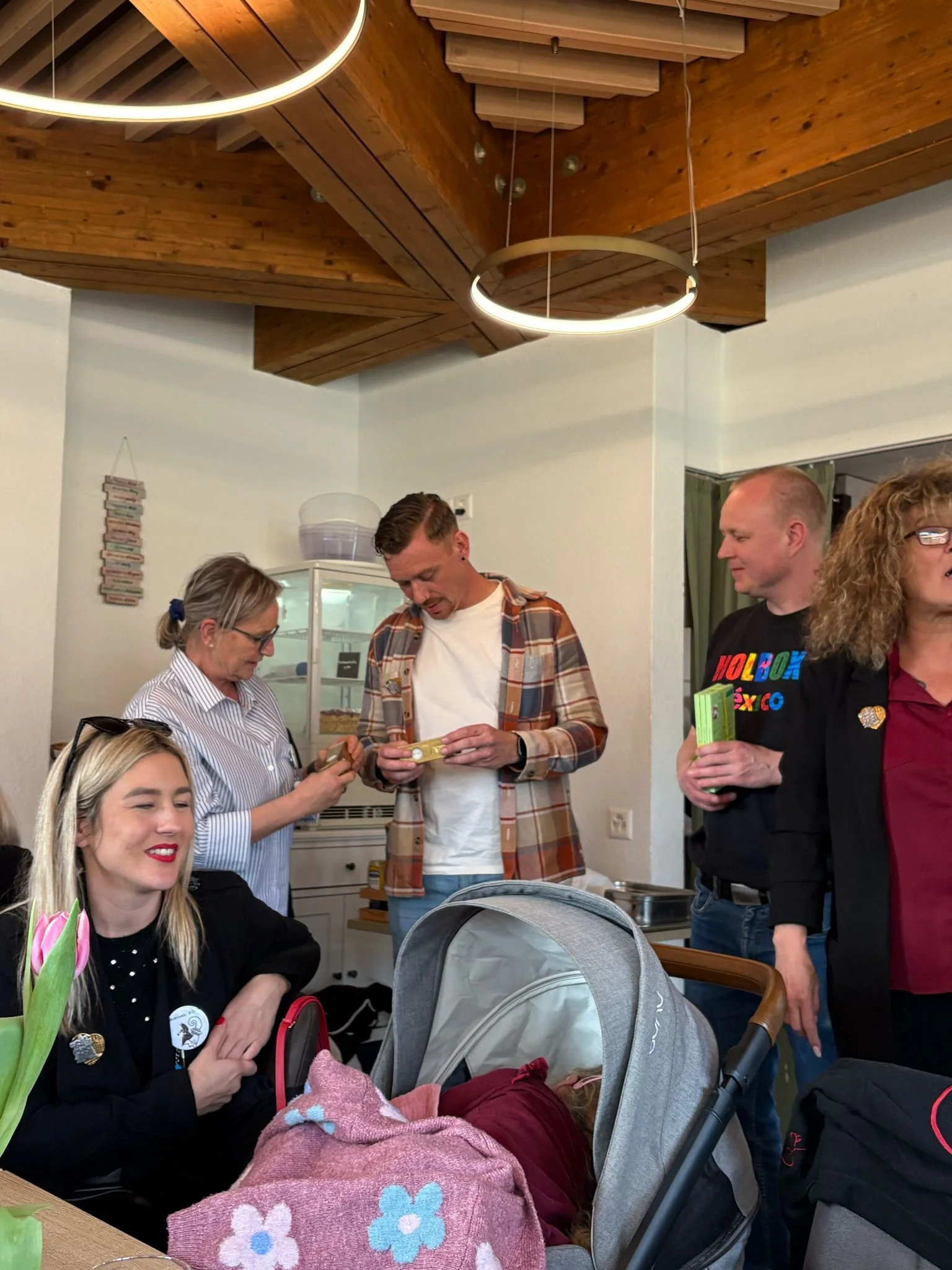 Mehrere Menschen, darunter eine Frau im Vordergrund mit einem Baby im Kinderwagen, in einem gemütlichen Raum mit Holzdecke, scheinen eine Zusammenkunft zu haben.