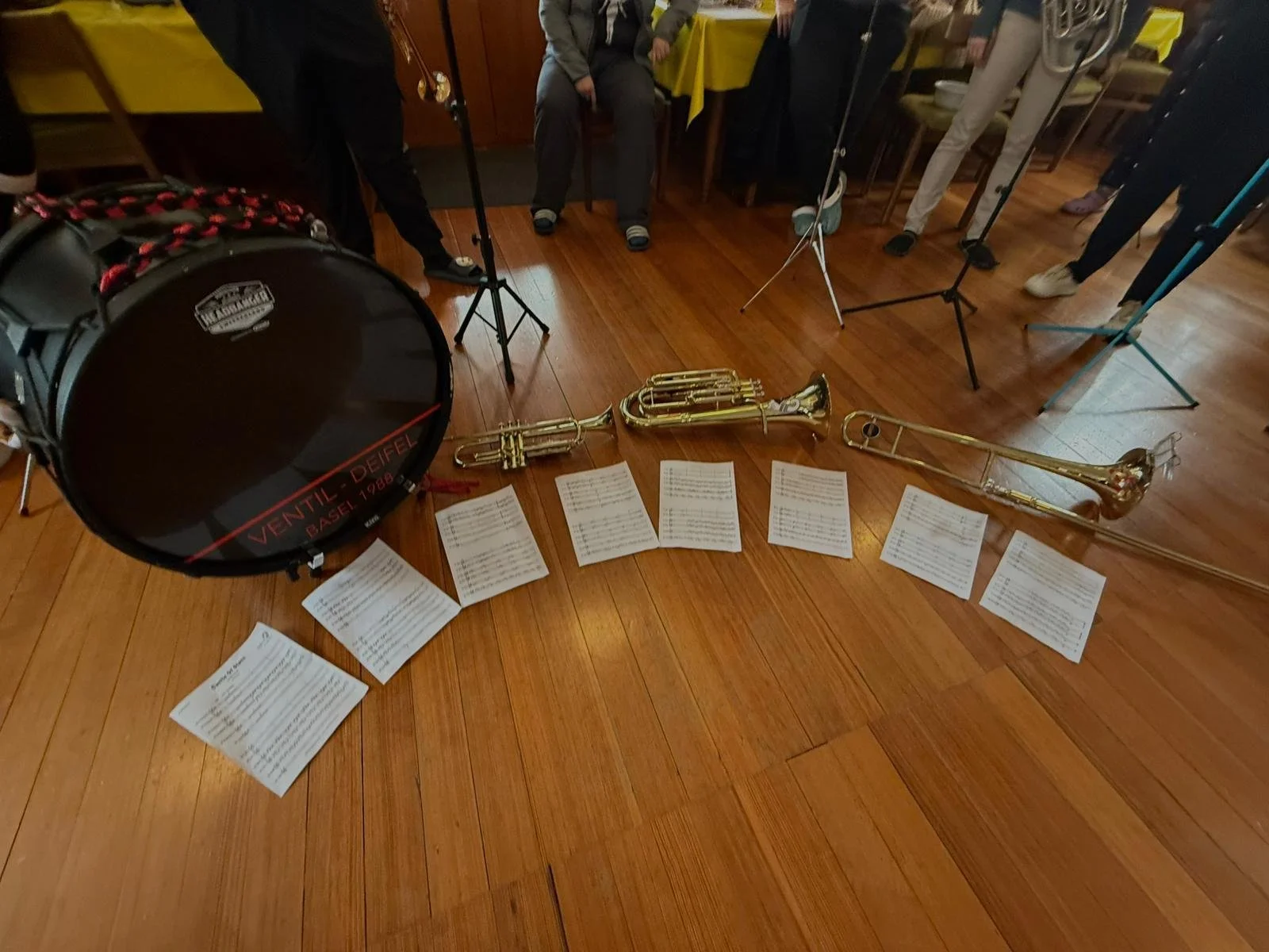 Bläserensemble mit Snare-Drum, Saxophon, Trompete, und Posaune auf Holzboden, Notenblätter liegen vor den Instrumenten, im Hintergrund Musiker und Musikzubehör.