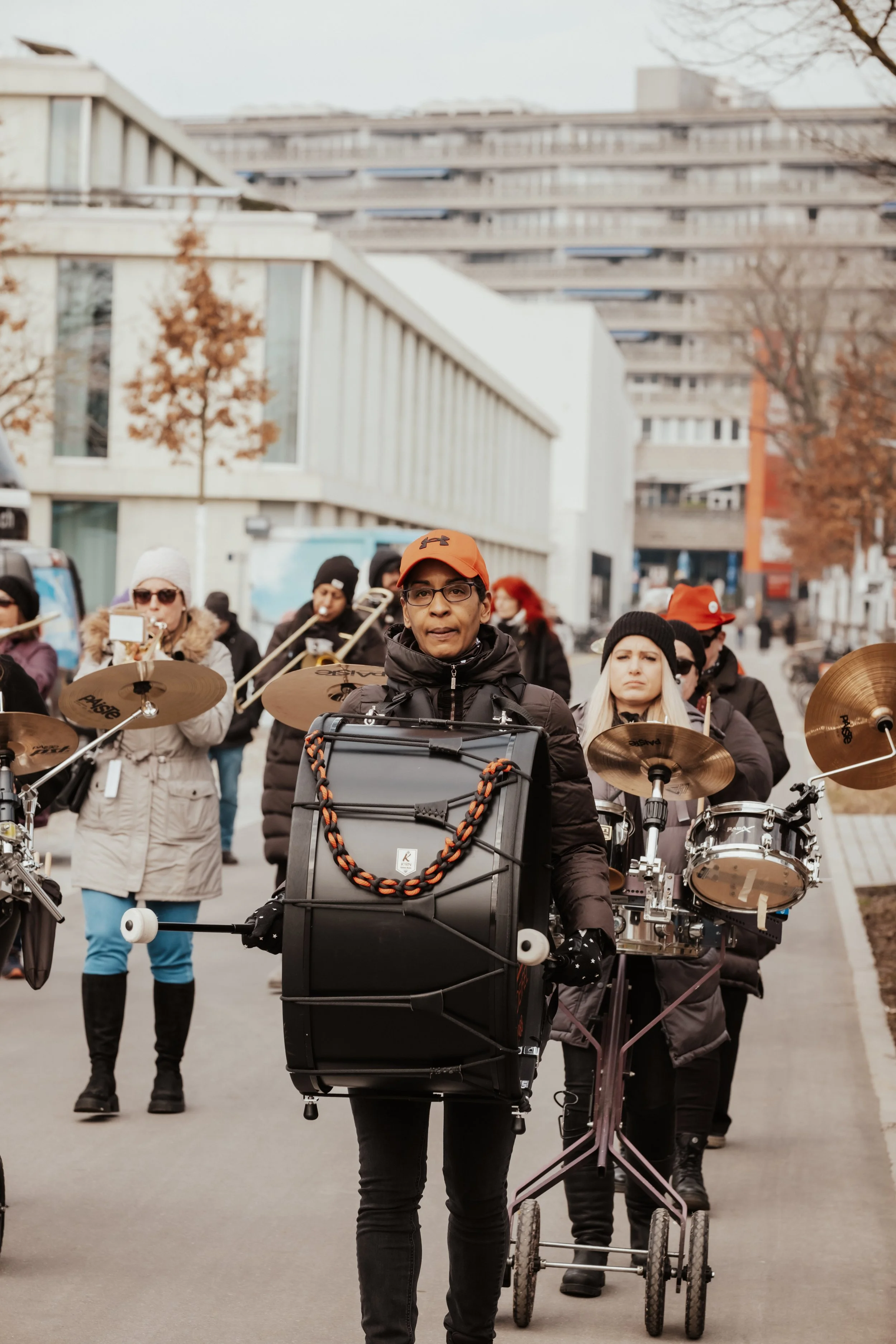 Gruppe von Musikern, die an einem Straßenkorso teilnehmen, mit verschiedenen Instrumenten, im Hintergrund moderne Gebäude und Bäume im Herbst.