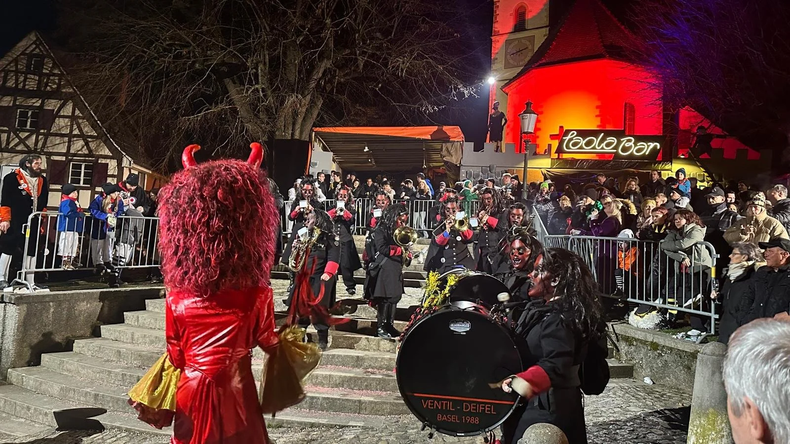 Menschen in Kostümen bei einer nächtlichen Parade oder einem Fest auf einer Straße, im Vordergrund ein Teufelskostüm mit roten Hörnern, im Hintergrund eine Gruppe von Musikern mit schwarzen Uniformen und rot akzentuierten Ärmeln, die auf Partituren u