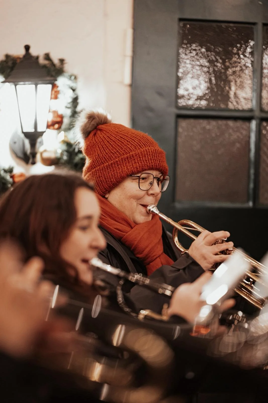 Zwei Frauen, eine mit roten Haaren und die andere mit Brille und rotem Mütze, spielen Blasinstrumente bei einer festlichen Veranstaltung.