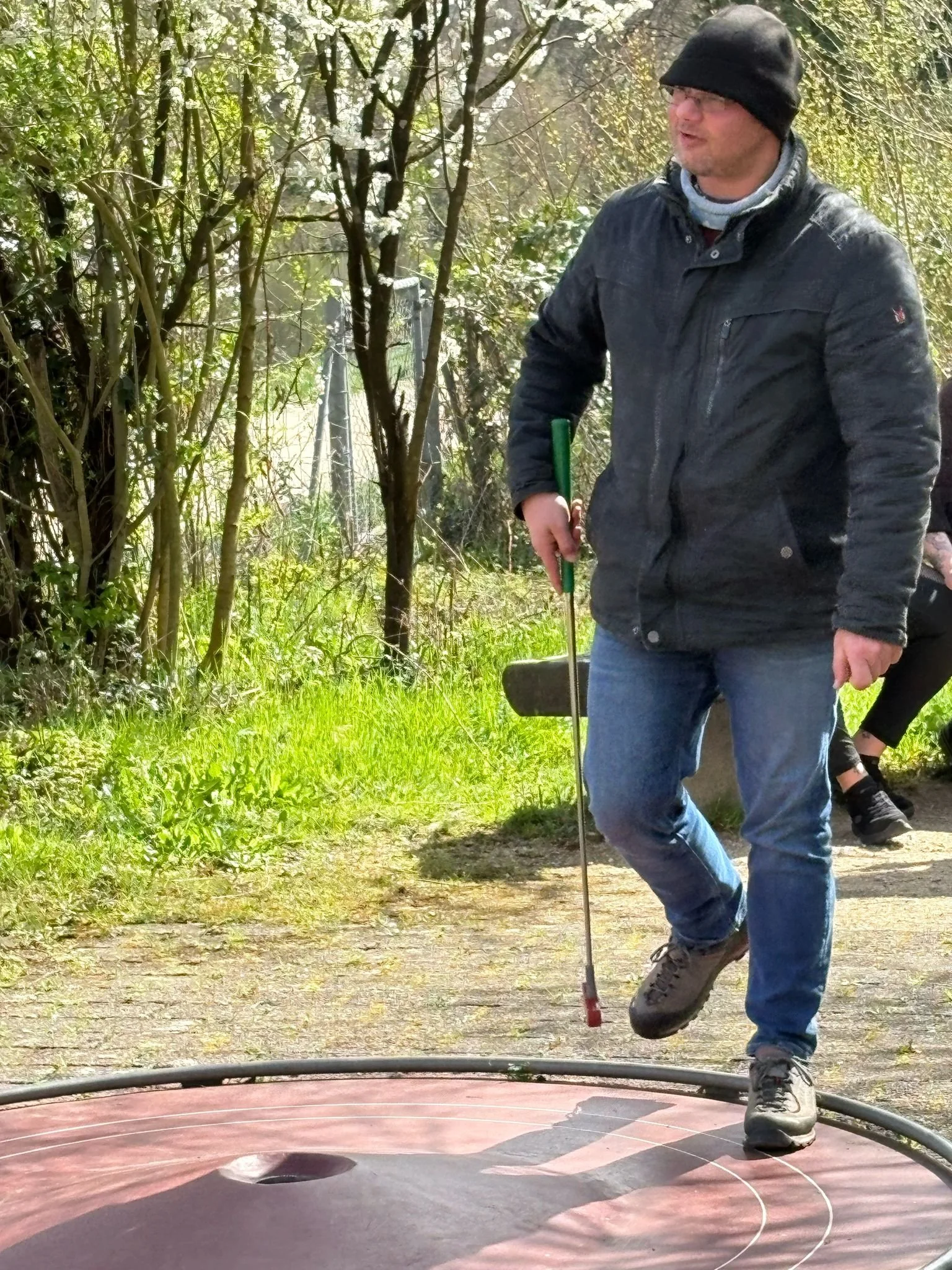 Mann beim Sporttraining auf einer blauen Matte im Freien, umgeben von Büschen und Bäumen, trägt dunkle Jacke, Mütze und Jeans, hält Stock in der Hand.