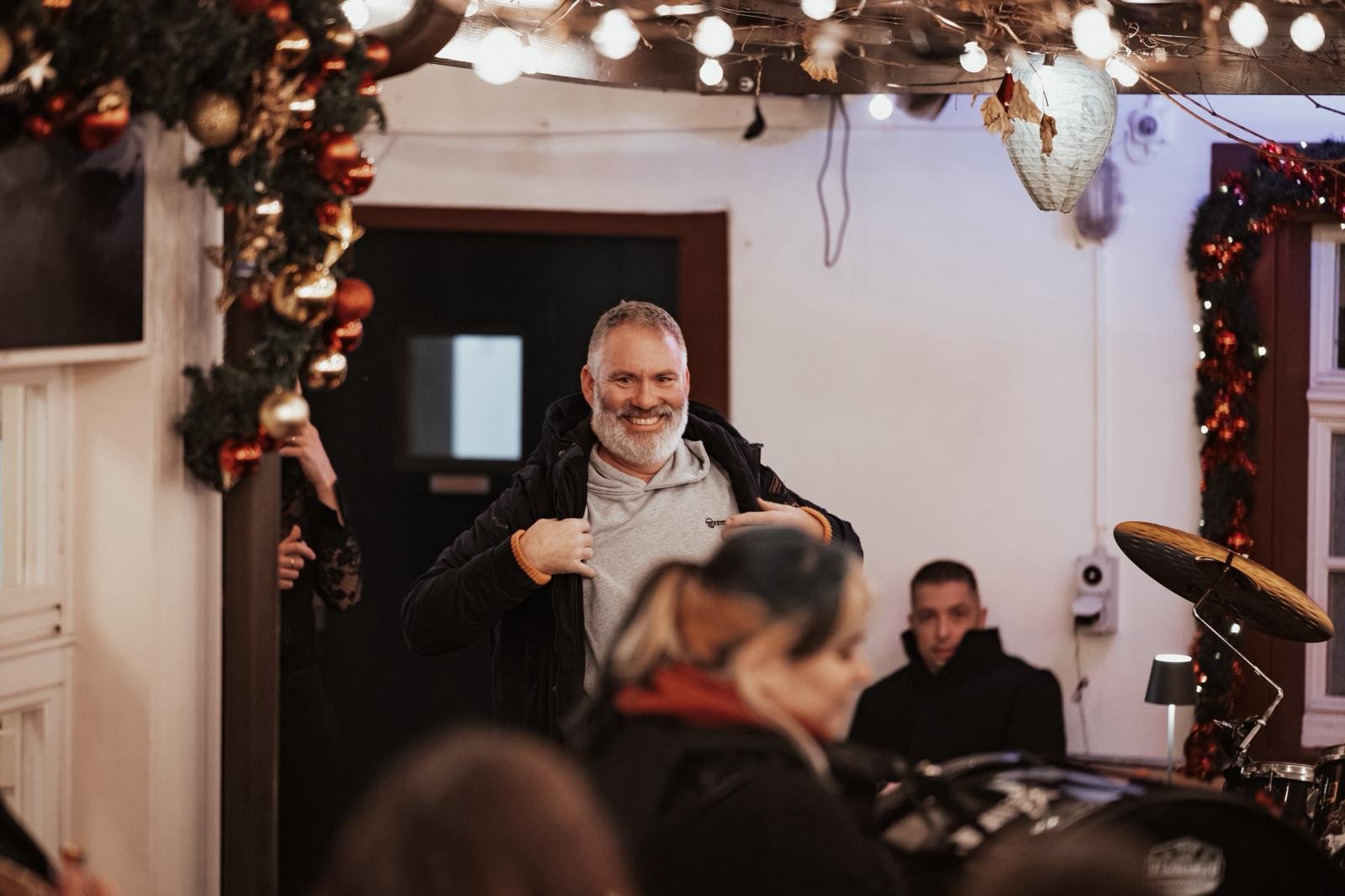 Ein Mann mit grauem Bart, der eine dunkle Jacke trägt, steht in einem festlich geschmückten Raum und lächelt. Im Hintergrund sind Weihnachtsdekorationen wie Kugeln und Lichter zu sehen, sowie andere Personen, die sitzen oder stehen.