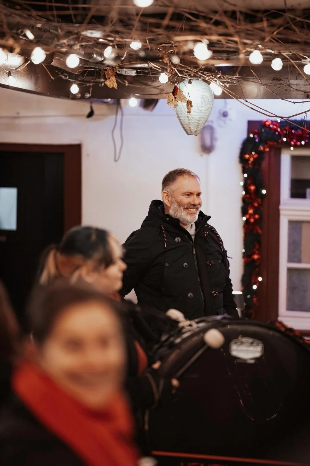 Ein smiling alter Mann steht in einer festlich geschmückten Raum mit Weihnachtsdekoration und Lichtern, umgeben von mehreren Menschen, die sich auf einem festlichen Event befinden.