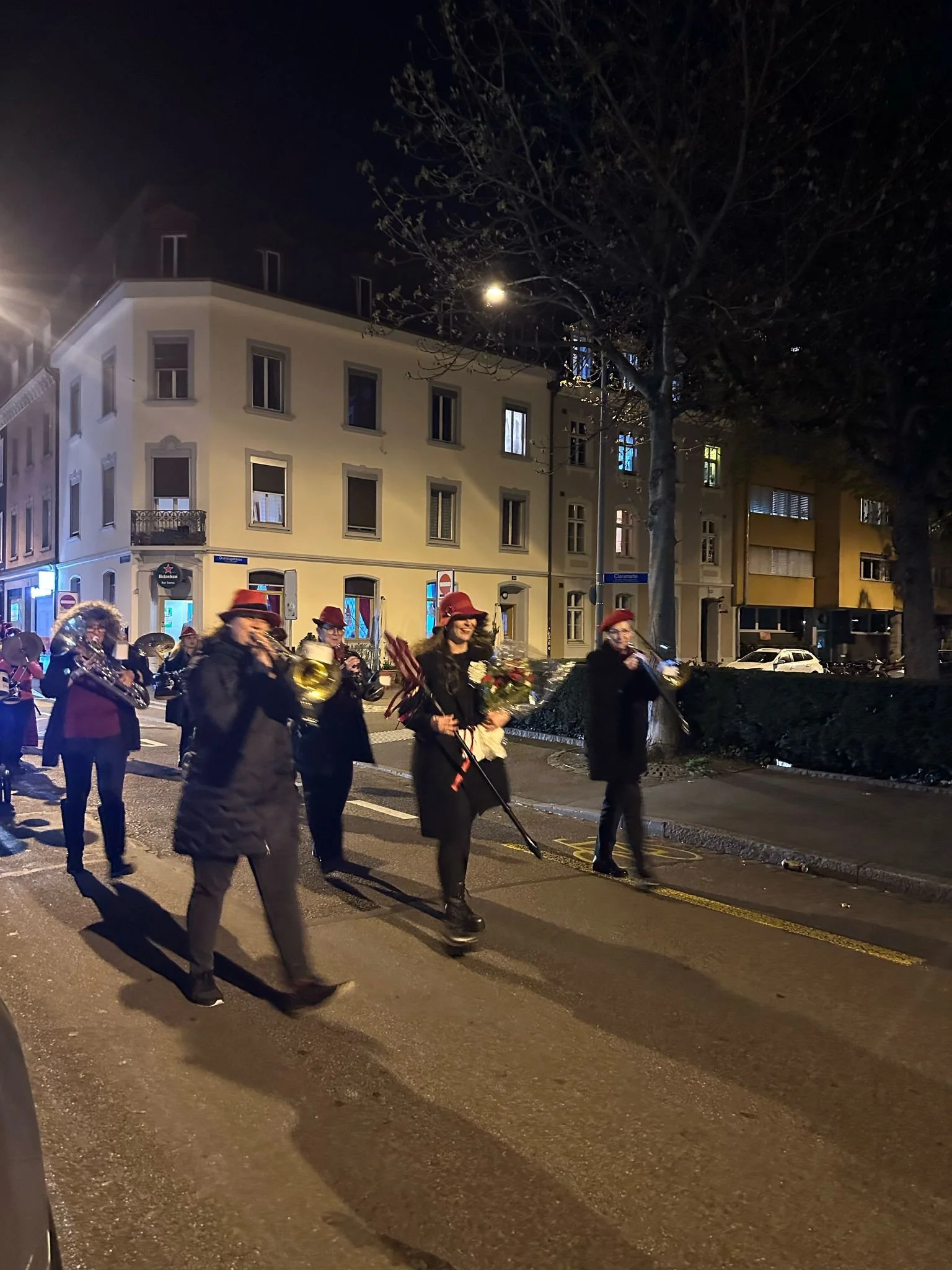 Eine Gruppe von Musikern, die nachts in einer Stadt auf der Straße marschieren. Sie tragen rote Hüte und spielen verschiedene Instrumente. Eine Frau in der Mitte hält einen Blumenstrauß. Im Hintergrund sind mehr Gebäude und Bäume bei Dunkelheit zu se