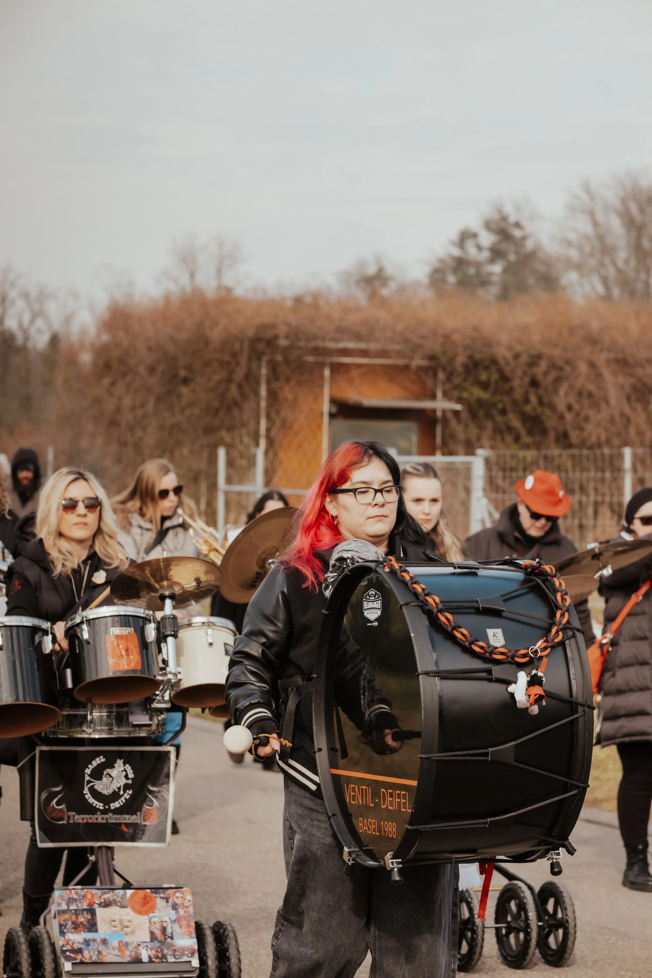 Eine Gruppe von Menschen mit Musikinstrumenten, darunter eine Frau mit roten Haaren, die eine große Tambourine spielt, während andere im Hintergrund auf einem offenen Gebiet musizieren.