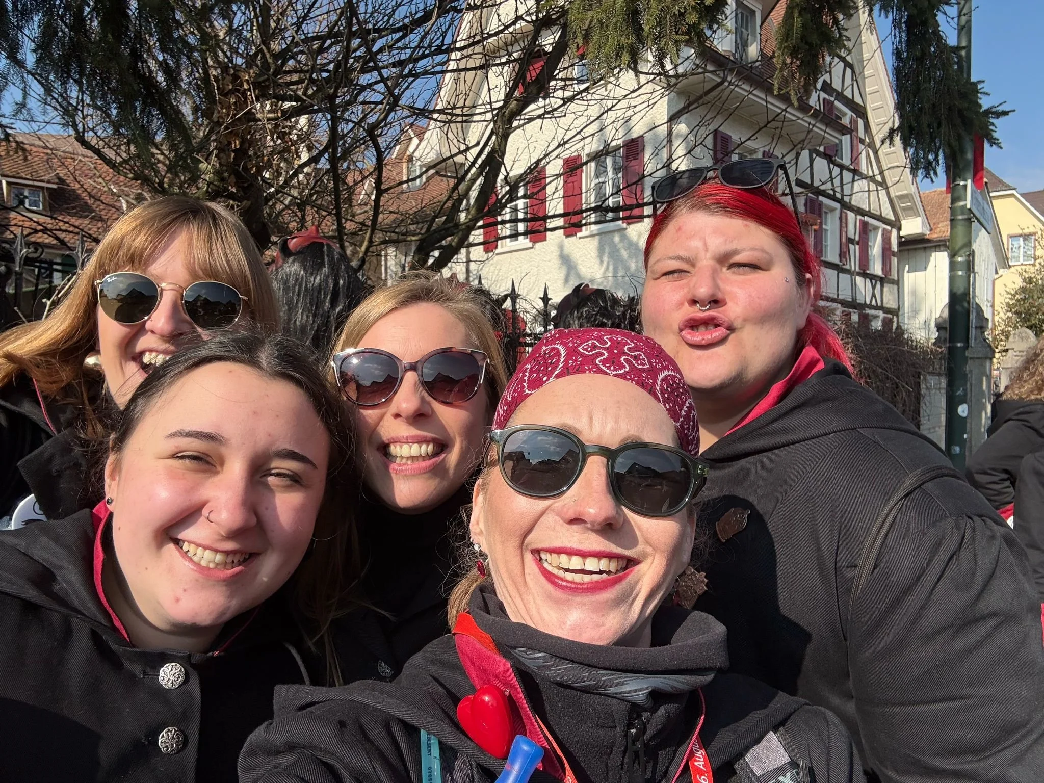 Mehrere lachende Frauen fotografieren sich für ein Selfie im Freien bei sonnigem Wetter, mit traditionellen europäischen Häusern im Hintergrund.