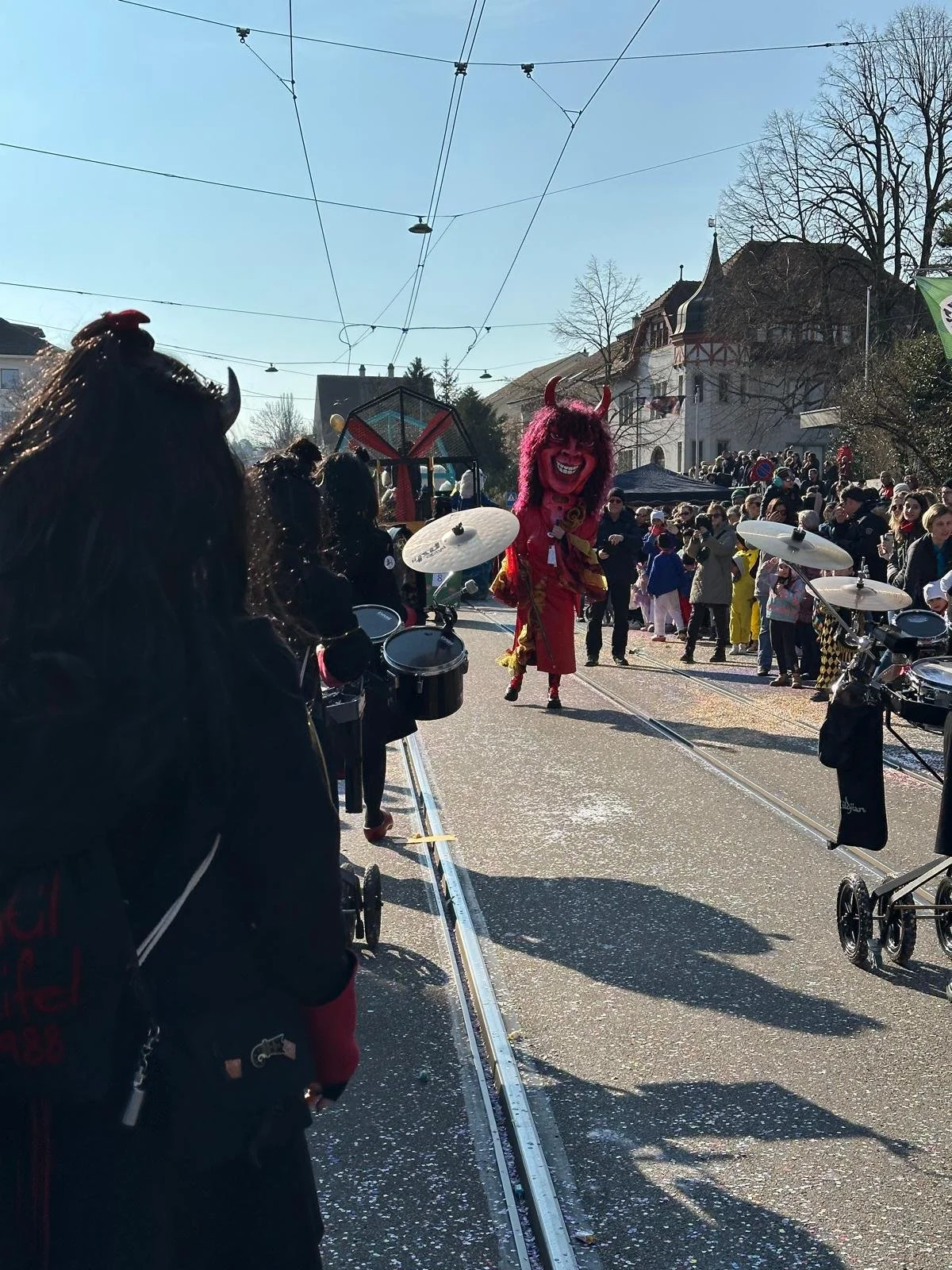 Menschen feiern Karneval im Freien, ein verkleideter Teufel tanzt in der Mitte, im Hintergrund stehen viele Zuschauer, sonniger Himmel, Trambahnschienen auf der Straße