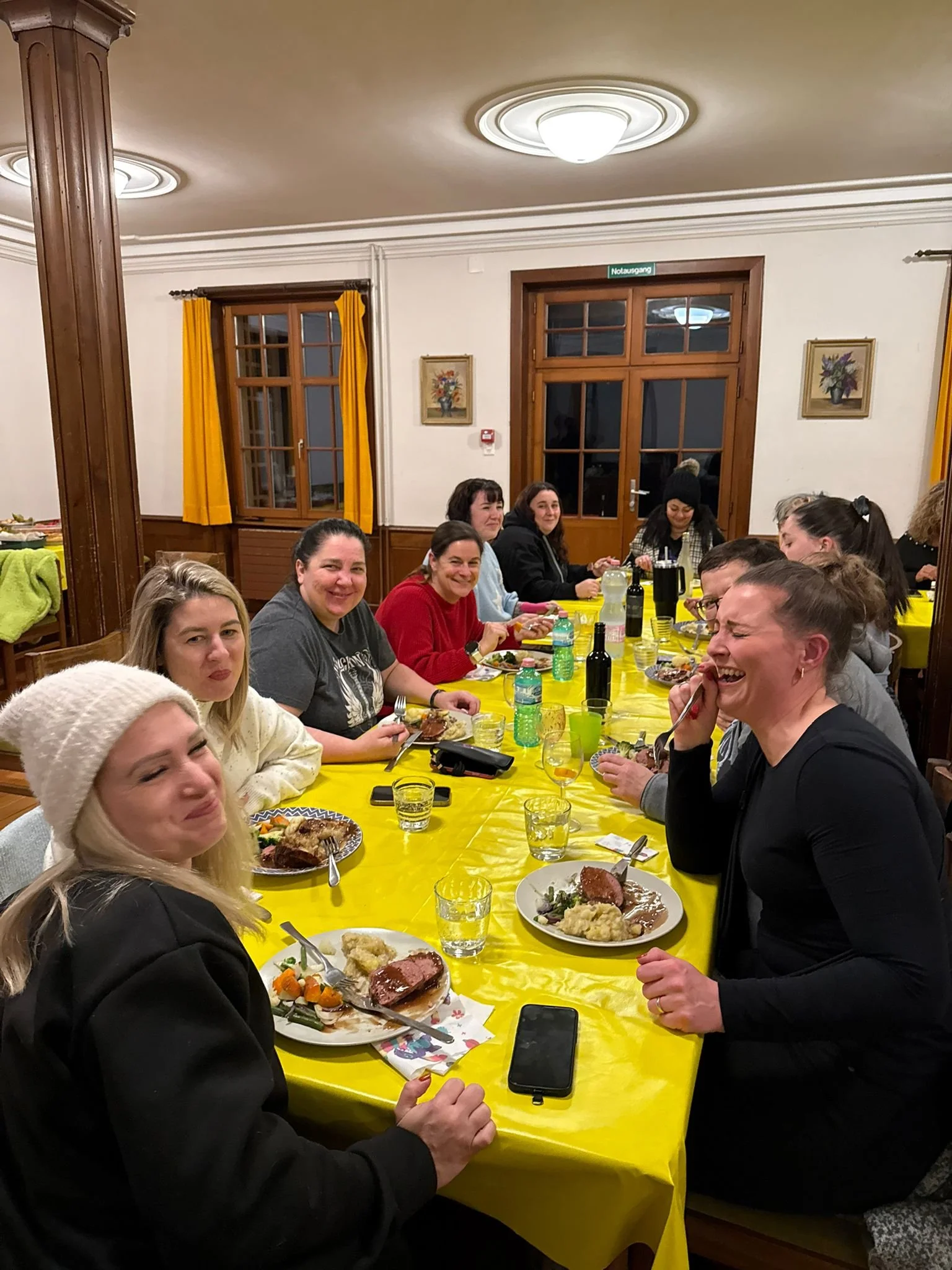Eine Gruppe von Menschen sitzt an einem langen Tisch mit gelber Tischdecke, isst und lacht in einem festlich dekorierten Raum.