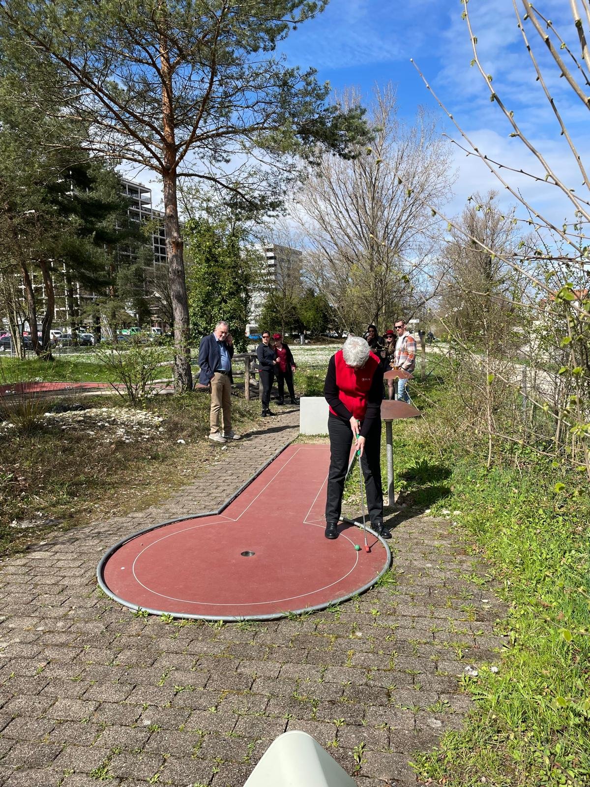 Menschen spielen Minigolf im Freien an einem sonnigen Tag, mit Bäumen und Gebäuden im Hintergrund.