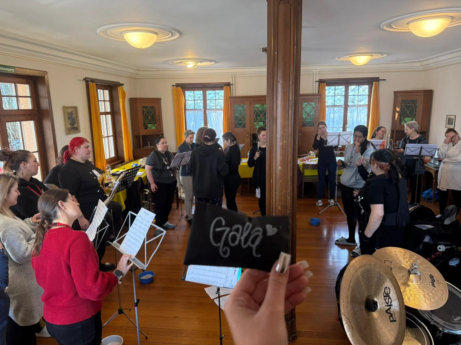 Gruppe von Frauen in einem gemütlichen Raum, die Proben mit Musikinstrumenten und Notenblättern machen, mit einer Hand, die ein Schild mit der Aufschrift "Gala" hält.