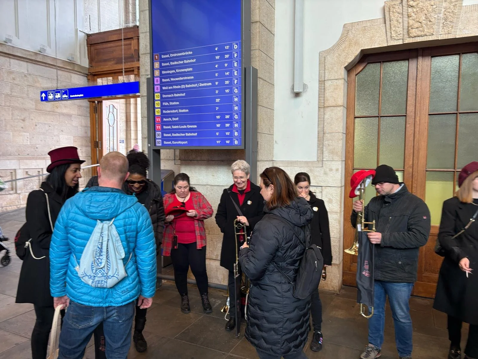 Menschen stehen in einer Bahnhofshalle, einige spielen Trompeten, andere schauen auf Mobiltelefone. Es gibt einen großen digitalen Anzeigetafel mit Abfahrtsinformationen und eine Beschilderung mit Richtungspfeilen.