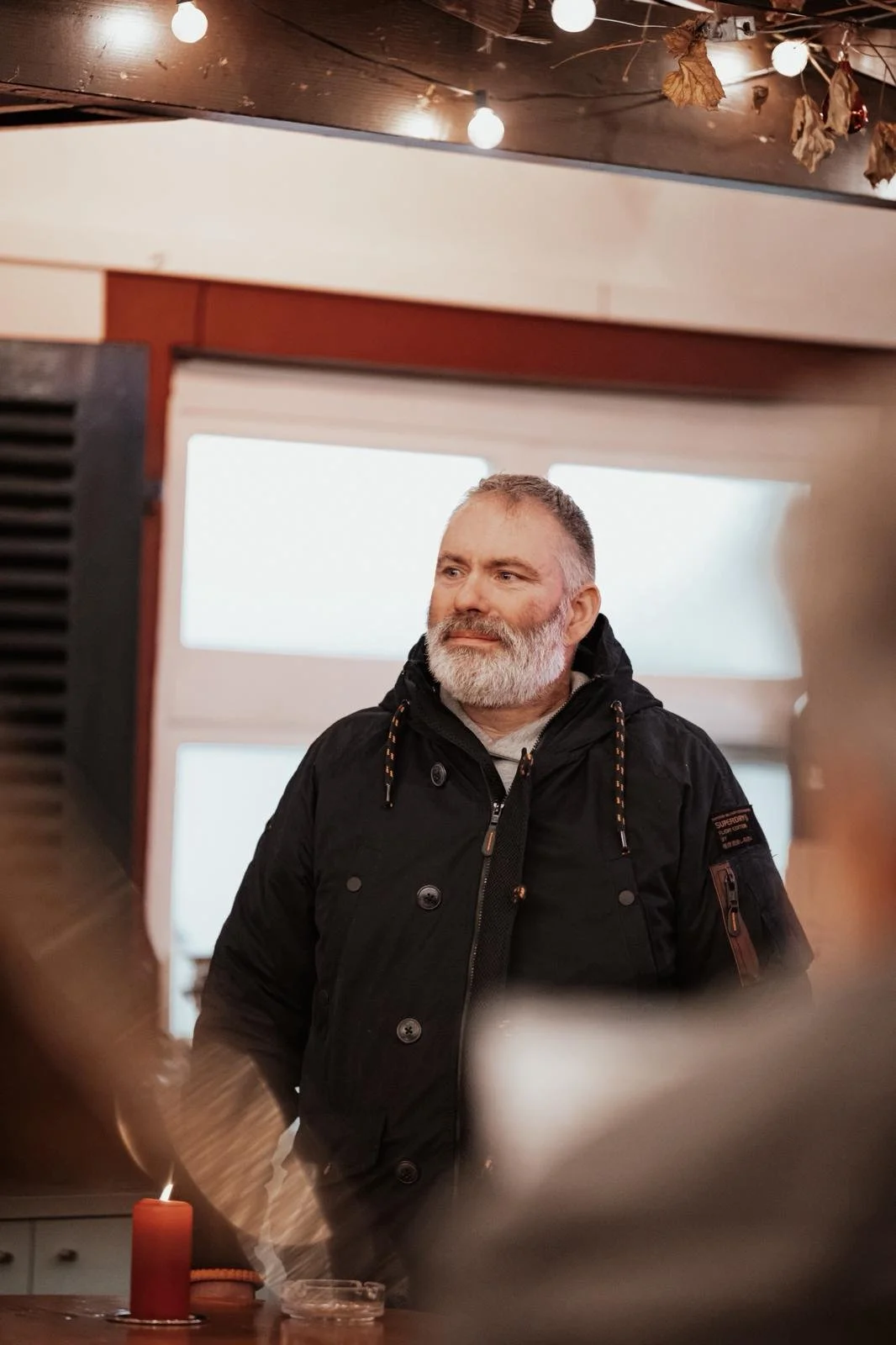 Ein älterer Mann mit grauem Bart, schwarzer Jacke, steht in einem Raum mit Fenstern im Hintergrund, dekoriert mit Lichterketten und getrockneten Pflanzen an der Decke.