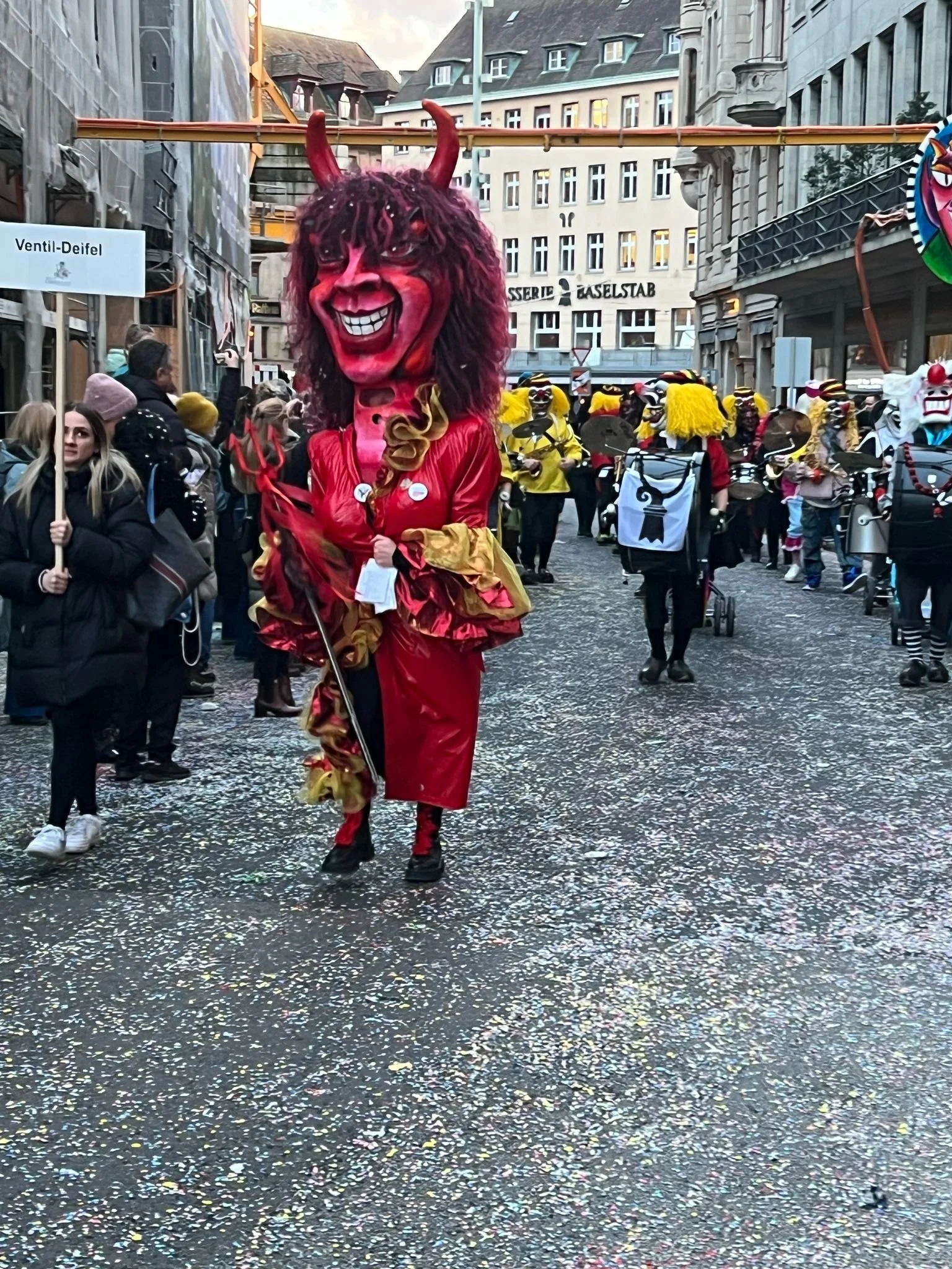 Eine Karnevalsfigur in einem Teufelskostüm mit großen roten Hörnern und einem großen Teufelsgesicht auf einem Umzug, umgeben von Menschen in festlicher Kleidung und weiteren Kostümen.