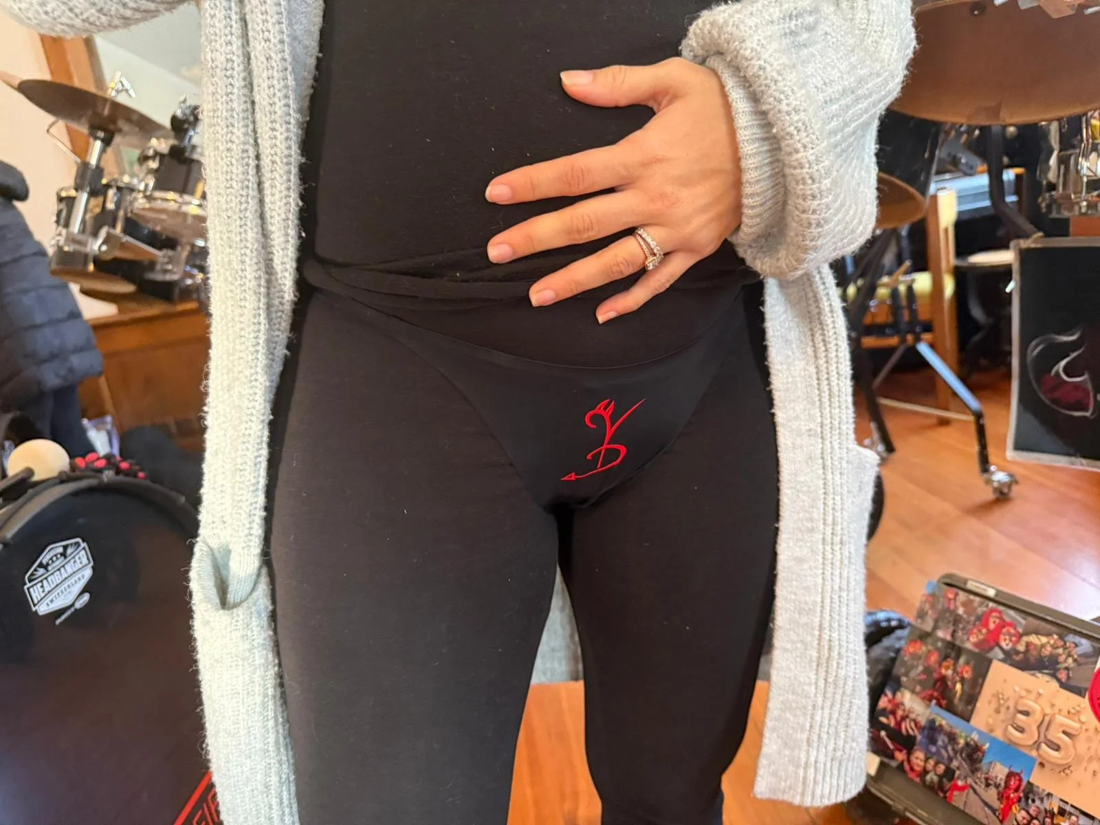 Nahaufnahme des unteren Teils eines Menschen in schwarzer Kleidung mit einer Hand, die auf den Bauch legt. Die Person trägt eine Ringe an einem Finger. Im Hintergrund sind Drehgeräte und andere Gegenstände sichtbar.