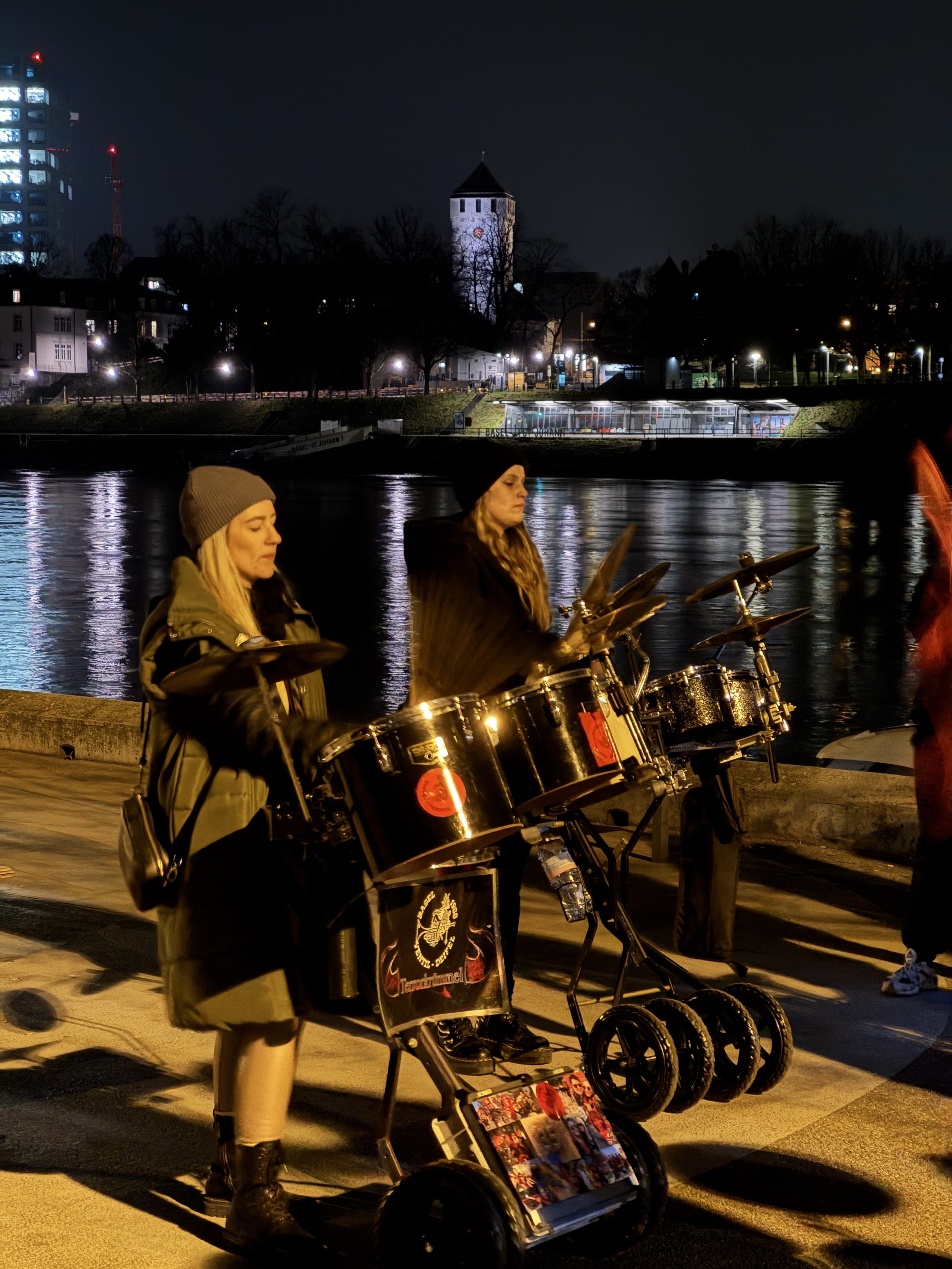 Zwei Straßenmusikerinnen mit Panflöten auf einem Stadtpromenadenabschnitt bei Nacht, im Hintergrund ein Fluss und eine beleuchtete Kirche