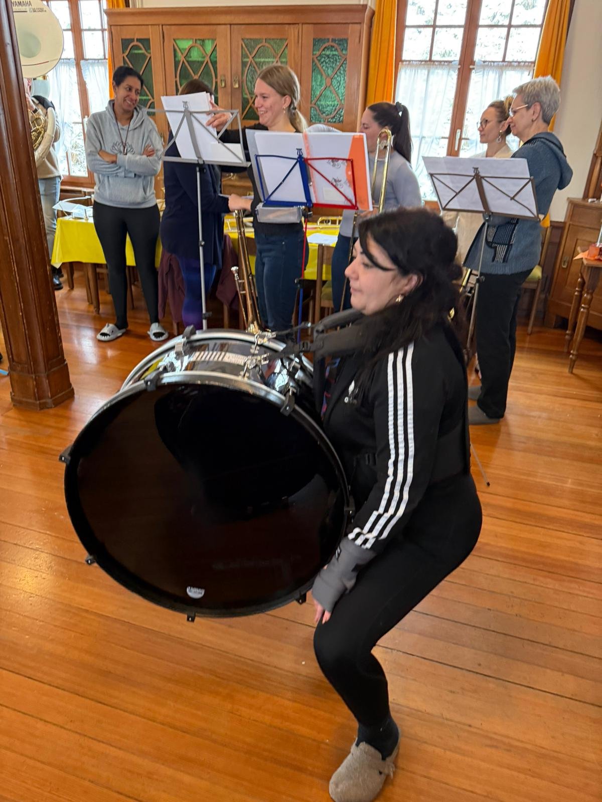 Eine Gruppe von Musikern, darunter eine Frau mit einer großen Trommel, die in einem Raum mit Holzboden und großen Fenstern spielt, während andere Musiker mit Notenständern im Hintergrund stehen.