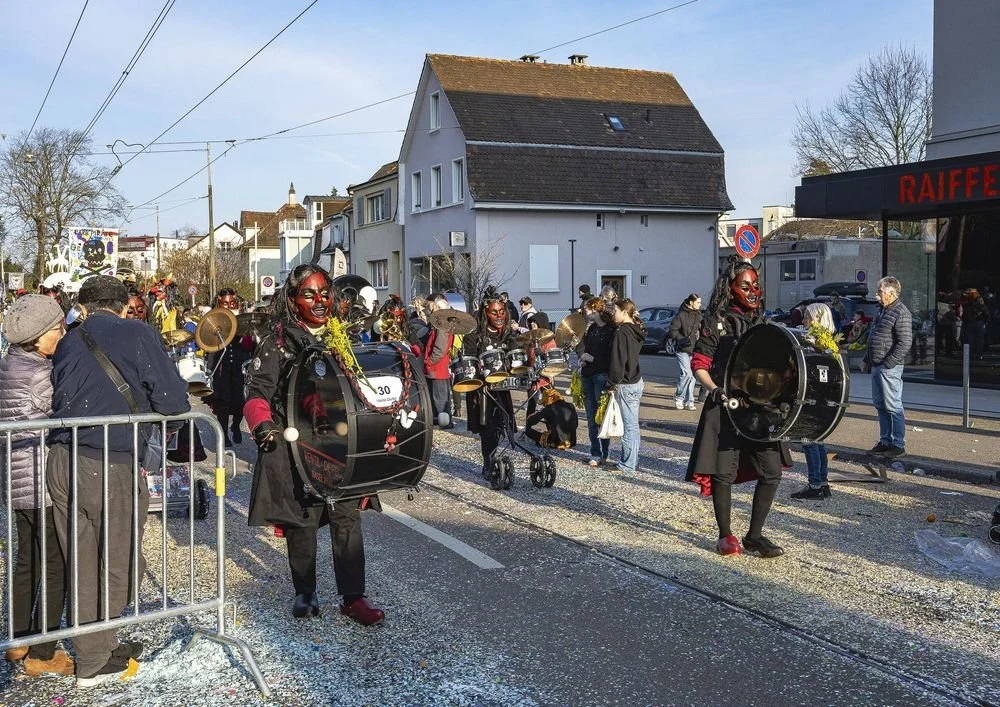 Menschen in einer Parade tragen schwarze Outfits und große Trommeln, während Zuschauer am Straßenrand zuschauen.