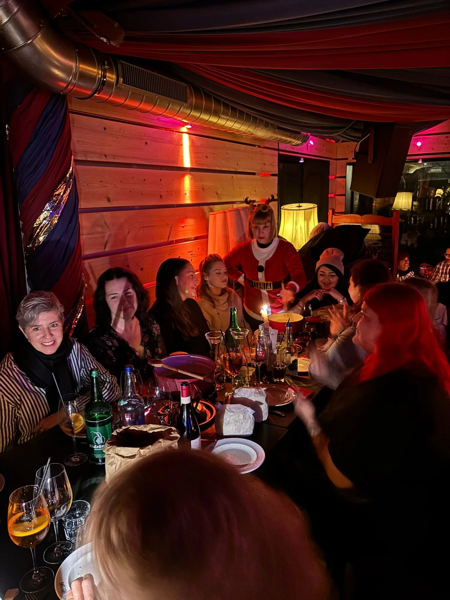 Mehrere Menschen sitzen an einem Tisch in einer festlich dekorierten Umgebung, während eine Person in einem roten Kostüm mit Rentiergeweihen eine Torte schneidet.