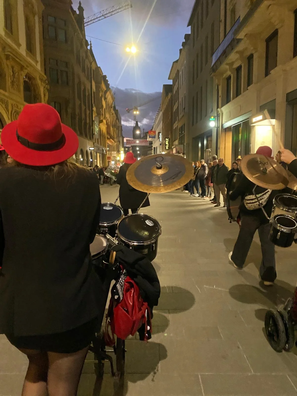 Menschen auf einer Stadtstraße bei Nacht, einige tragen rote Hüte, einige spielen Musikinstrumente, im Hintergrund erleuchtete Gebäude und ein Kirchturm.
