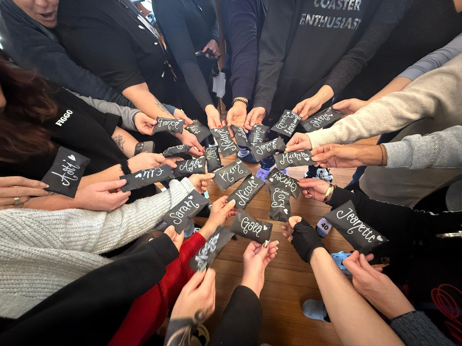 Mehrere Personen, die jeweils ein Stück schwarzen Pappe mit weißen Schreibschrift-Namen halten, die in einem Kreis auf einem Holztisch ausgerichtet sind.
