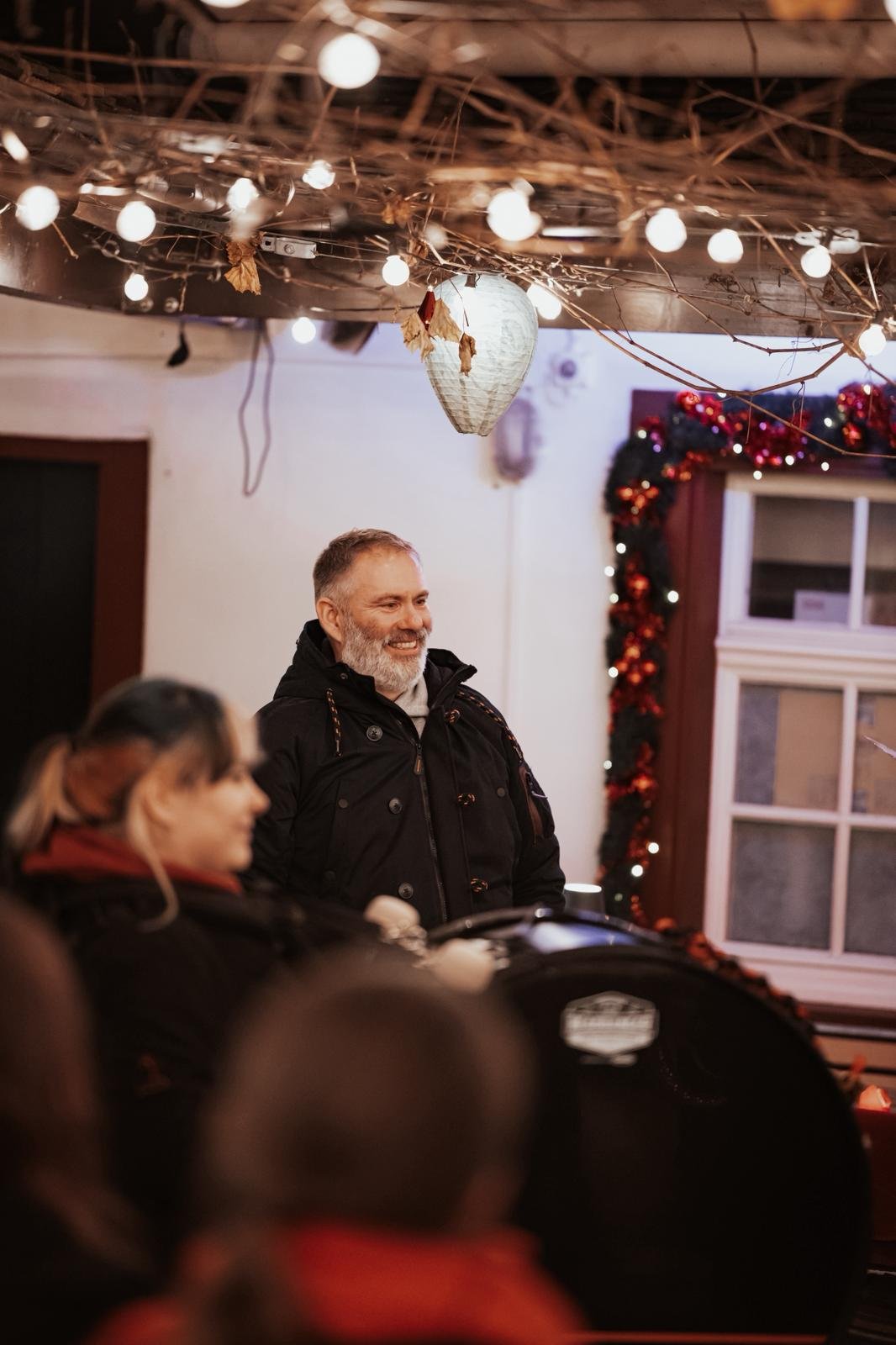Ein Mann mit grauem Bart steht lächelnd vor einer Gruppe von Menschen bei einer festlichen Veranstaltung. Im Hintergrund sind weihnachtliche Dekorationen wie Lichterketten, eine Weihnachtstanne mit roten Kugeln und ein Fenster zu sehen.