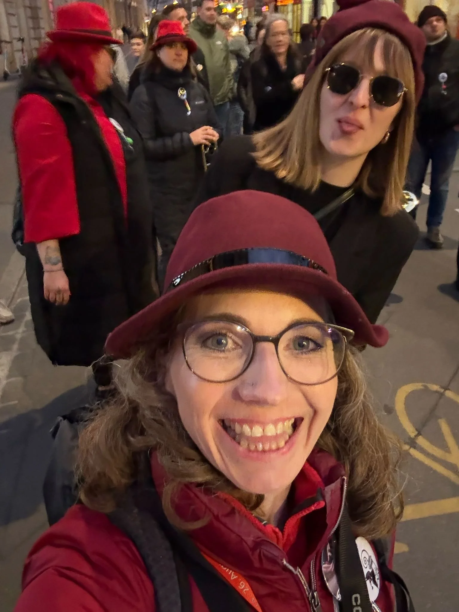 Zwei Frauen vor einer Menschenmenge auf der Straße, eine trägt eine rot-bordeauxfarbene Mütze und Brille, die andere eine schwarze Jacke, Sonnenbrille und eine Mütze, im Hintergrund stehen weitere Menschen, einige haben rote Kleidung, die Beleuchtung