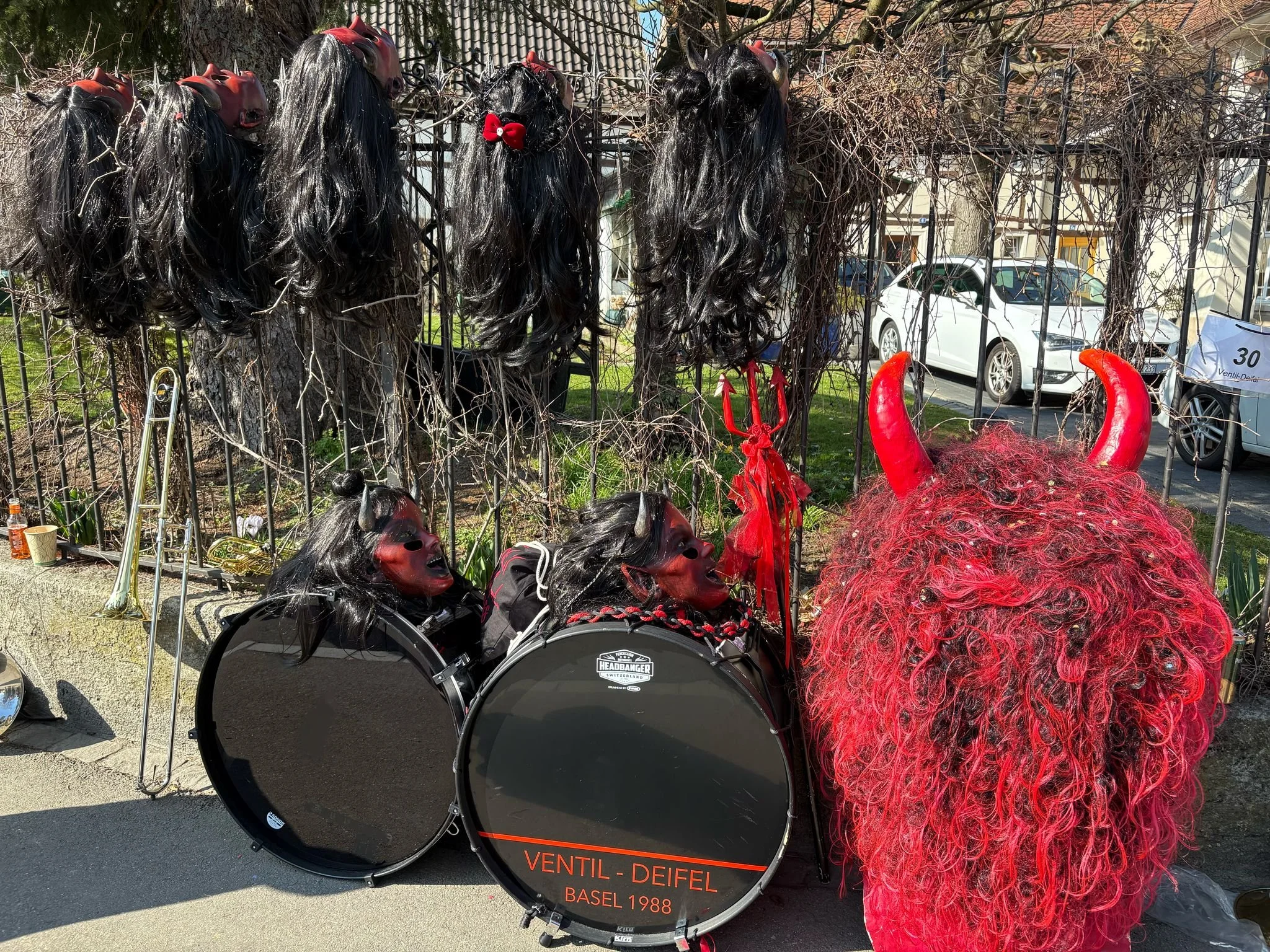 Kostüme und Masken, die an eine Teufelsfigur mit roten Hörnern und roter Schleier erinnern, liegen neben einem Schlagzeug, das mit Teufelsmasken dekoriert ist, und an einer Metallzaun hängen Perücken oder Masken mit schwarzen langem Haar und roten Ma