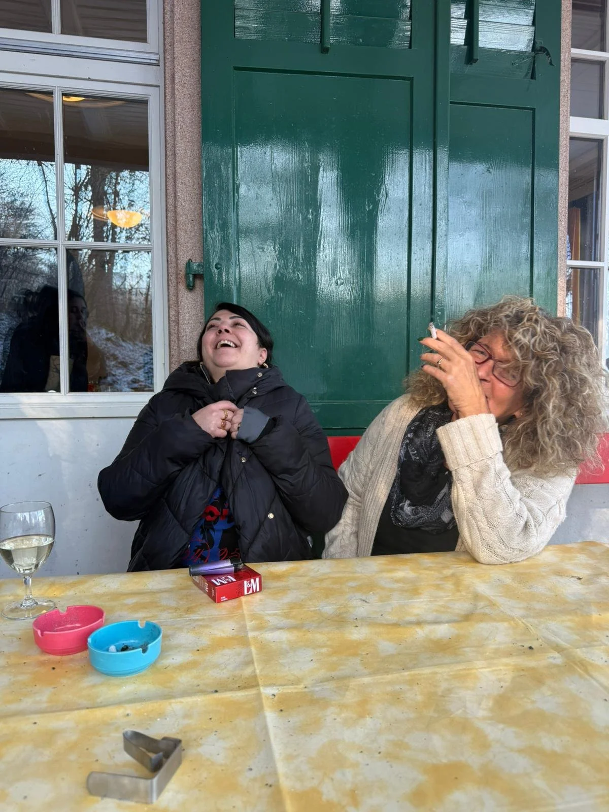Zwei lachende Frauen sitzen an einem Tisch, eine raucht Zigarette, auf dem Tisch liegen Zigaretten, Aschenbecher und ein Glas Wein im Vordergrund, im Hintergrund eine grüne Tür und Fenster, eine Person im Fenster schaut hinaus.