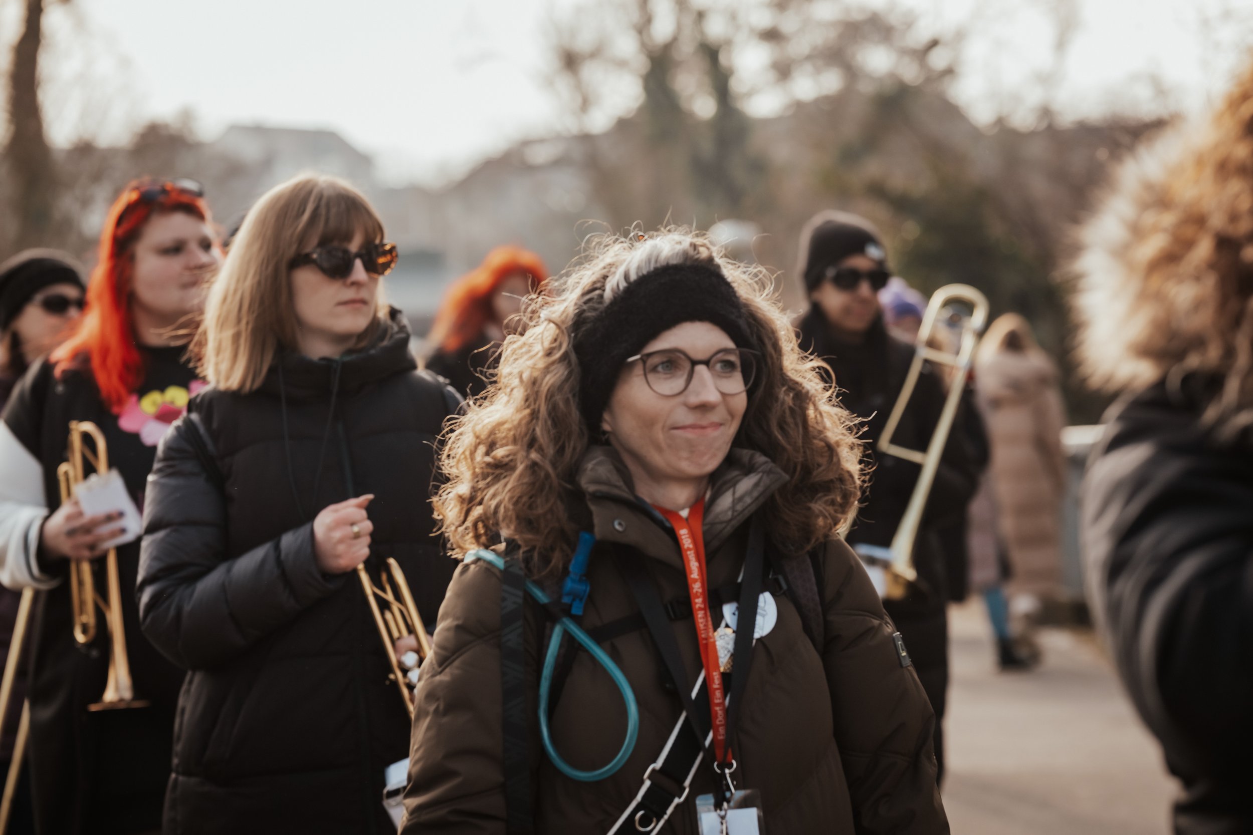 Eine Gruppe von Menschen, darunter eine Frau mit Locken, Brille und schwarzem Stirnband, die im Freien spazieren gehen, viele tragen dunkle Jacken und Sonnenbrillen, im Hintergrund sind Bäume und ein Fluss sichtbar.