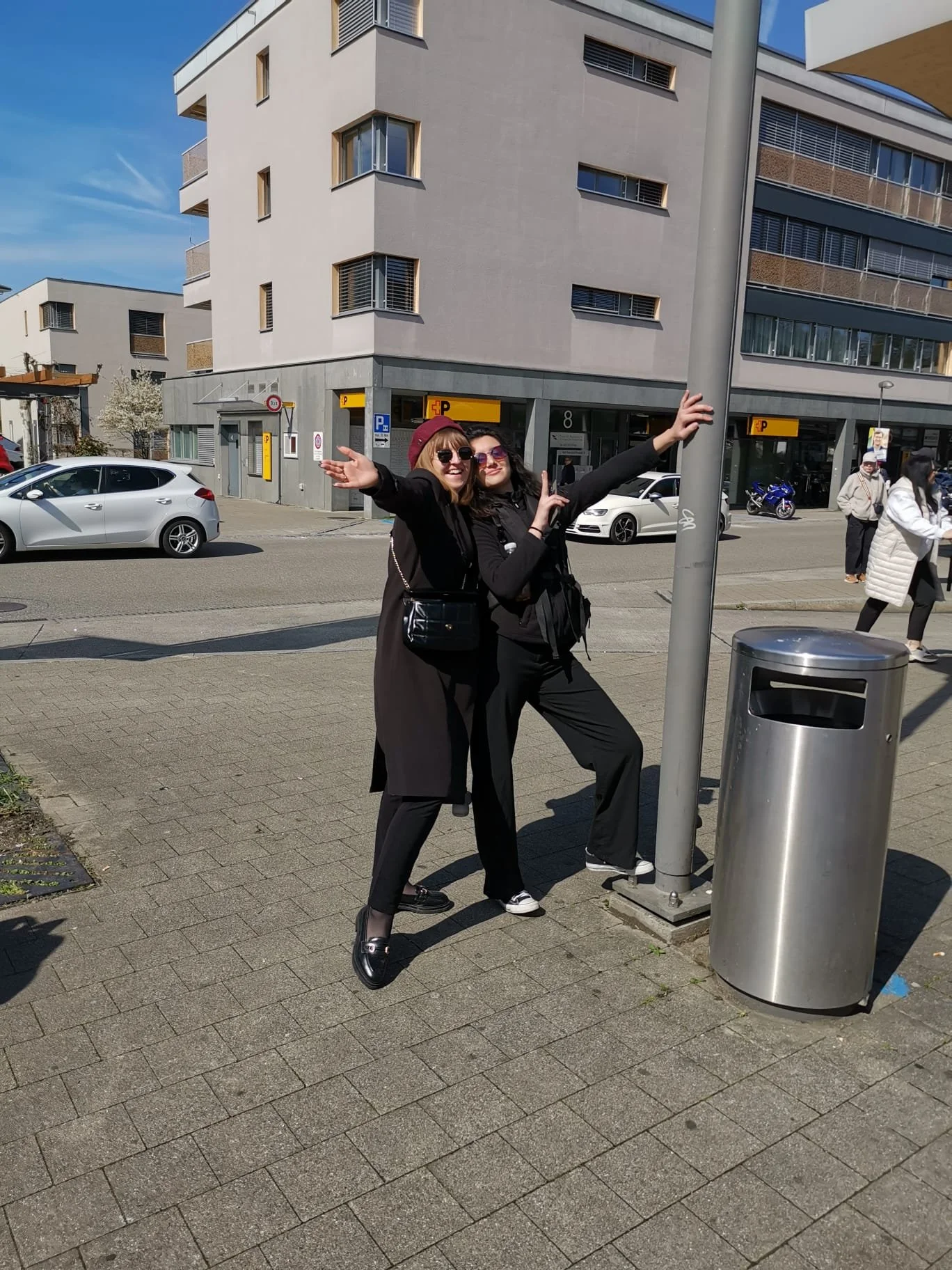 Zwei lachende Frauen stehen auf einem Bürgersteig, die eine mit ausgestreckten Armen, bei Sonnenschein, neben einem Laternenpfahl.