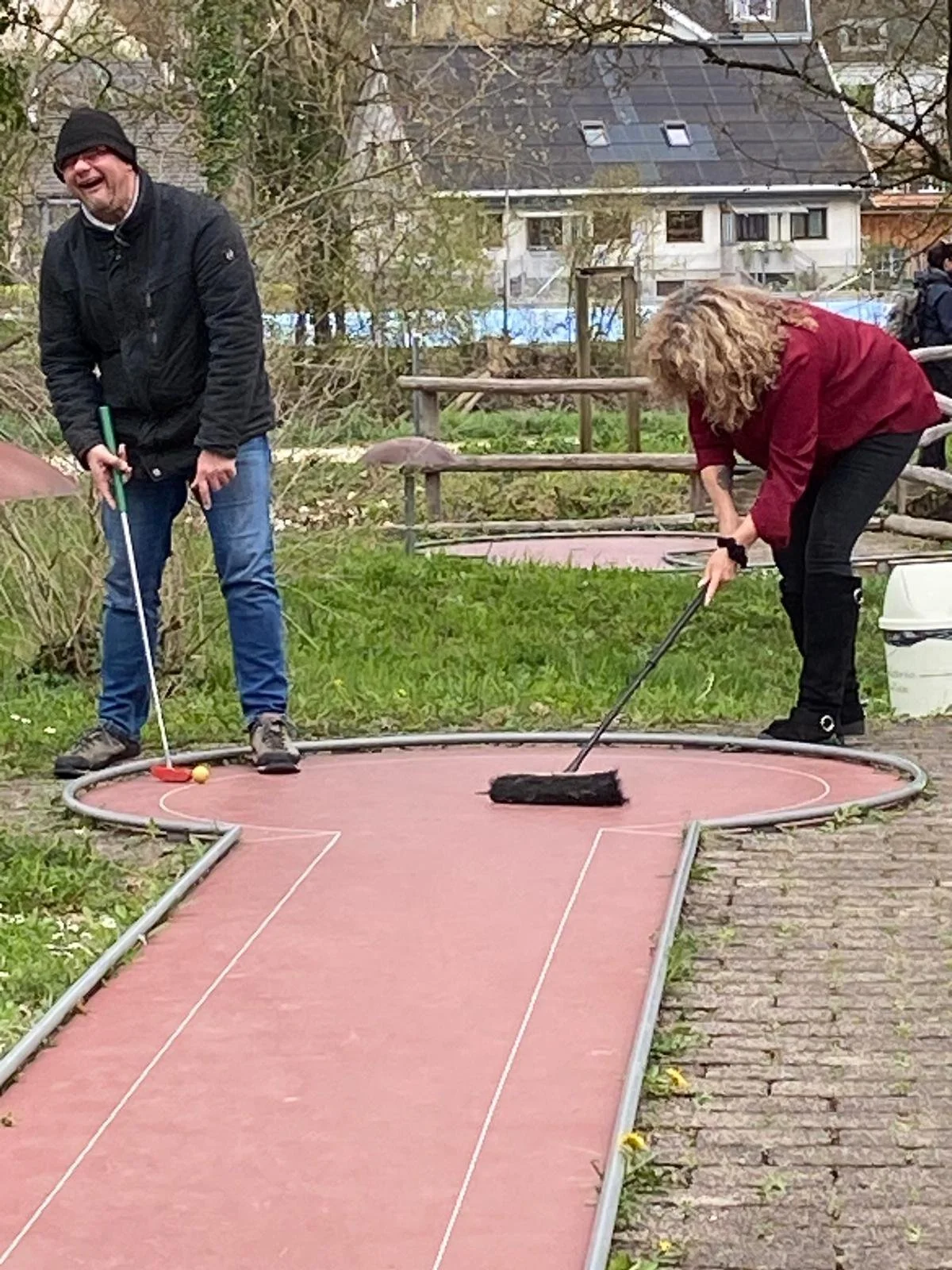 Zwei Personen spielen Mini-Golf im Freien. Einer schlägt den Ball, der andere reinigt die Minigolfbahn mit einem Besen. Es ist bewölkt, und die Umgebung ist grün mit Bäumen und Gebäuden im Hintergrund.