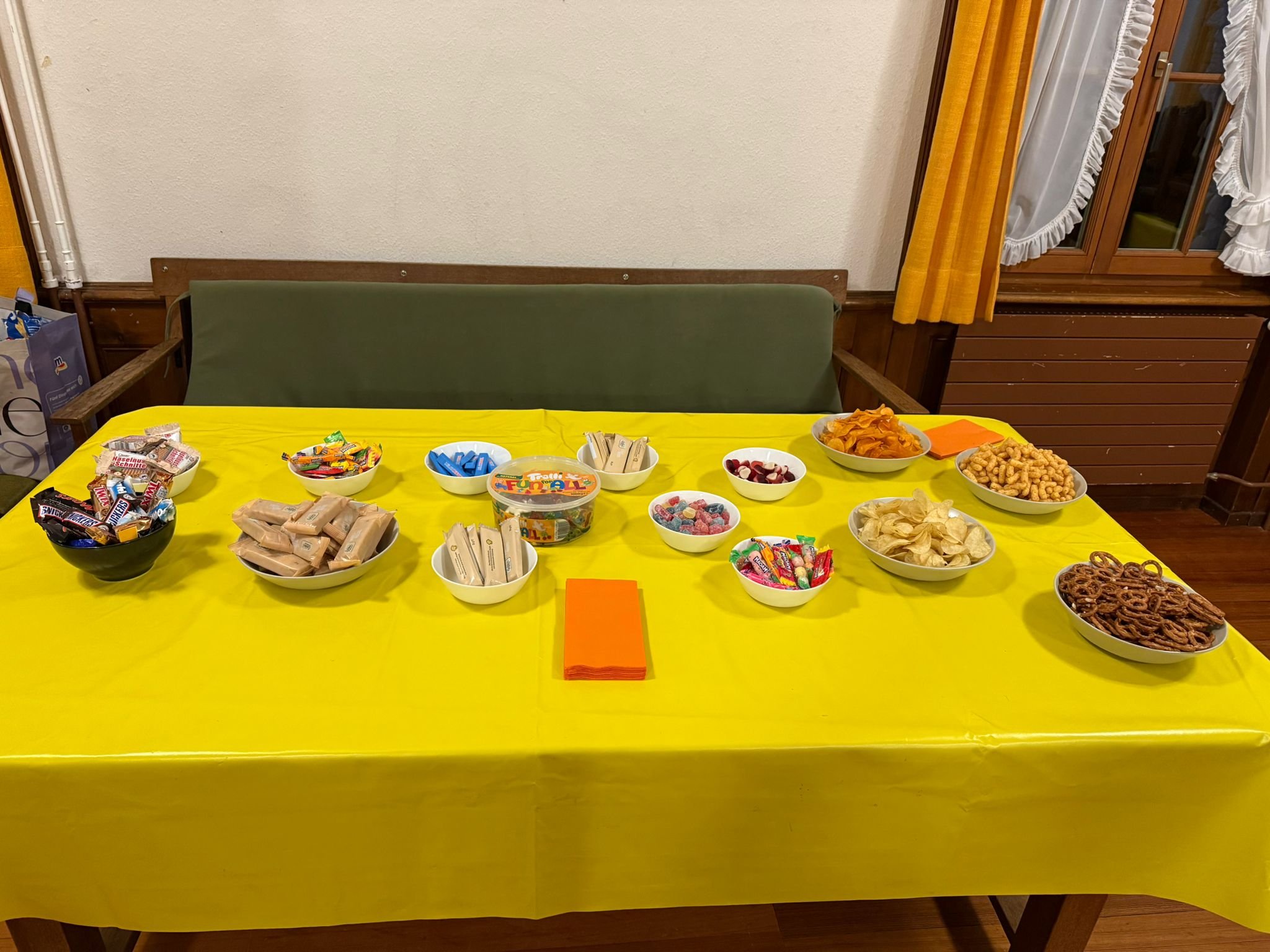 Tisch mit gelber Tischdecke, darauf verschiedene Süßigkeiten und Snacks in Schalen, im Hintergrund eine grüne Bank, Fenster mit weißen Vorhängen und einer gelben Gardine.