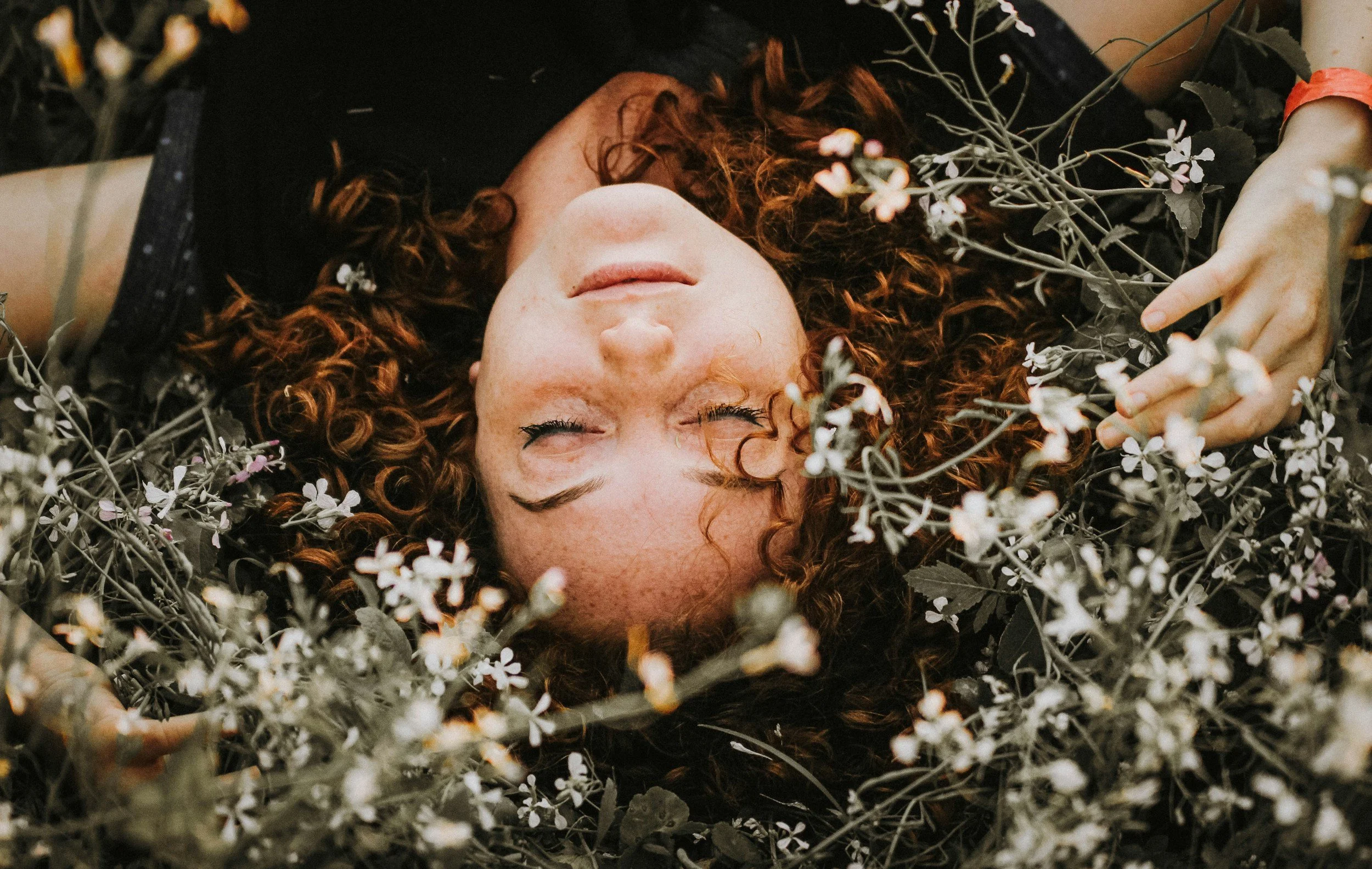 A woman with curly red hair lying on her back surrounded by small white flowers, her eyes closed and her face peaceful.