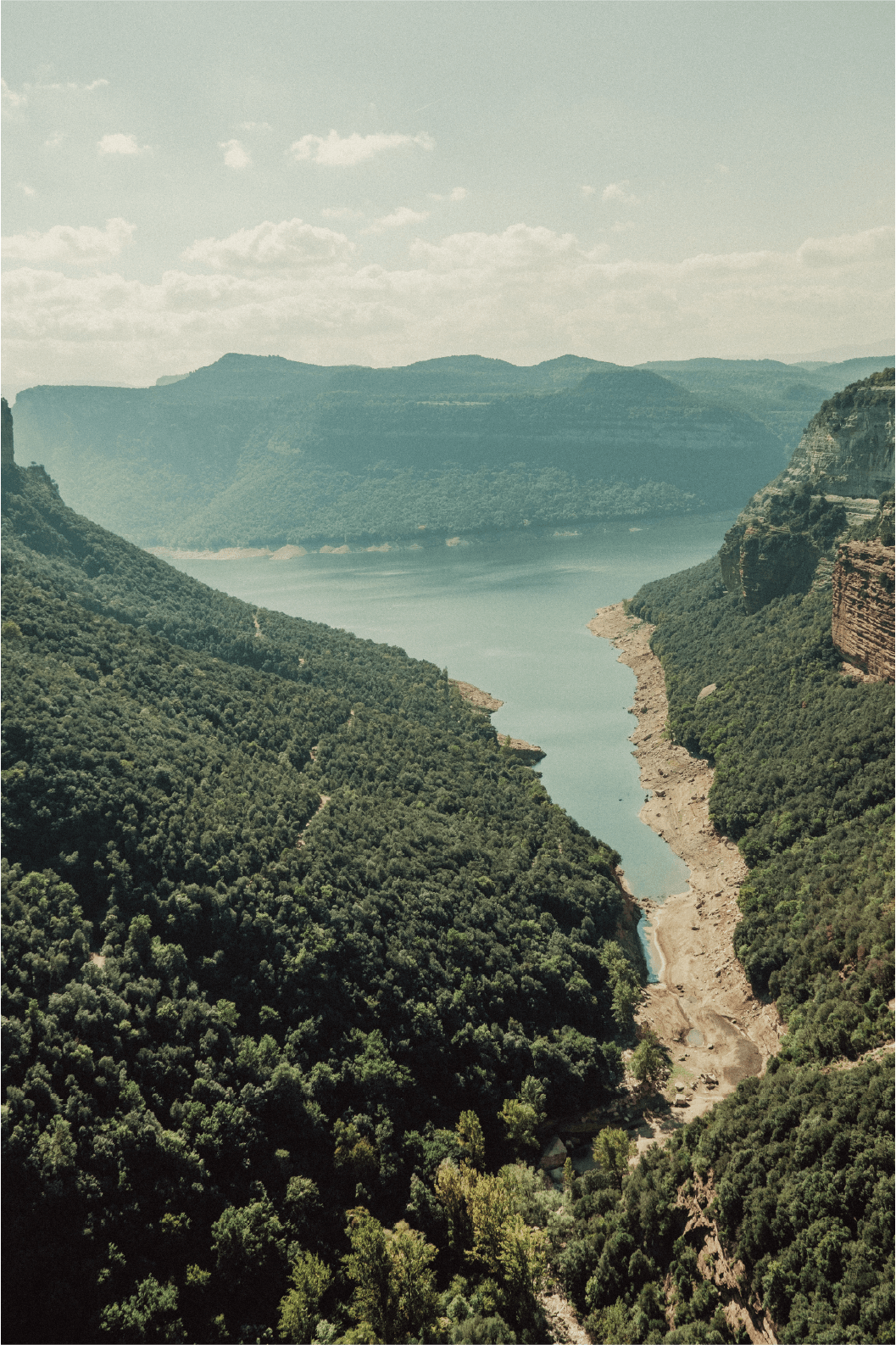 A scenic view of a river winding through a lush green canyon with forested hills and dramatic cliffs under a partly cloudy sky.