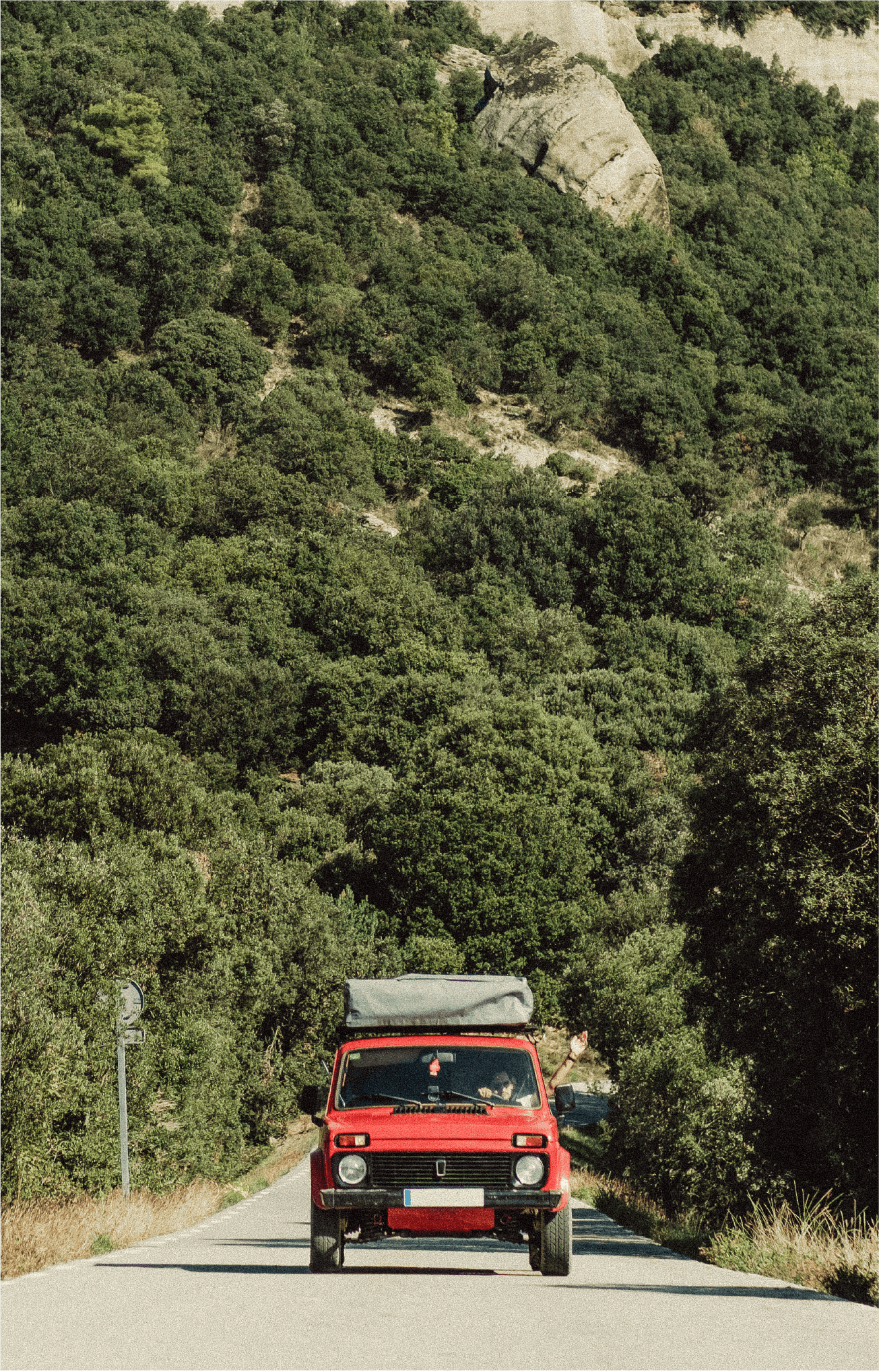 A red vintage car with a roof rack driving on a narrow road surrounded by dense green trees and hills, with a rocky outcrop visible in the background.