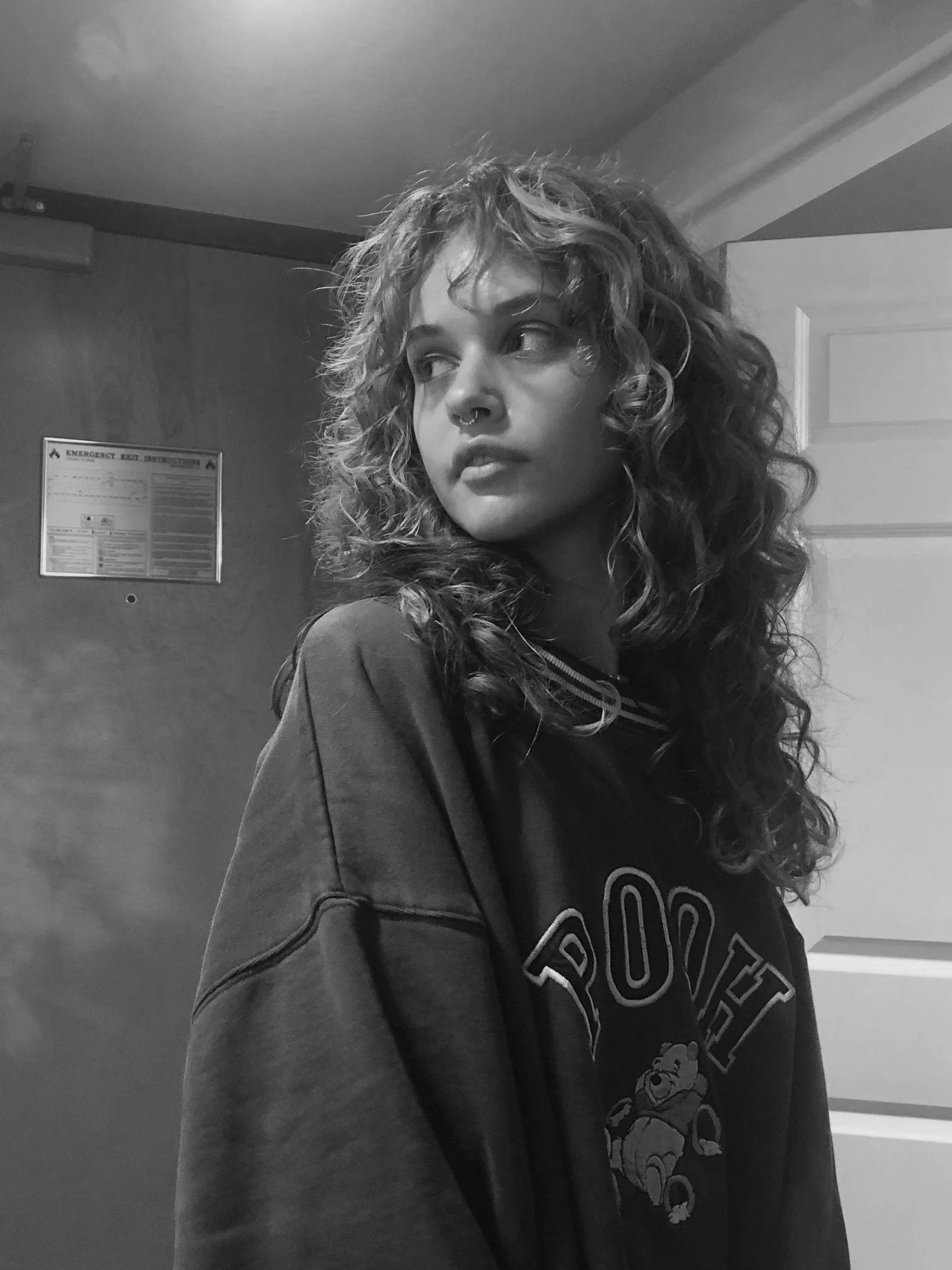 Black and white photo of a young woman with curly hair, wearing a sweatshirt, looking to her left, standing in front of a door with an emergency exit sign.