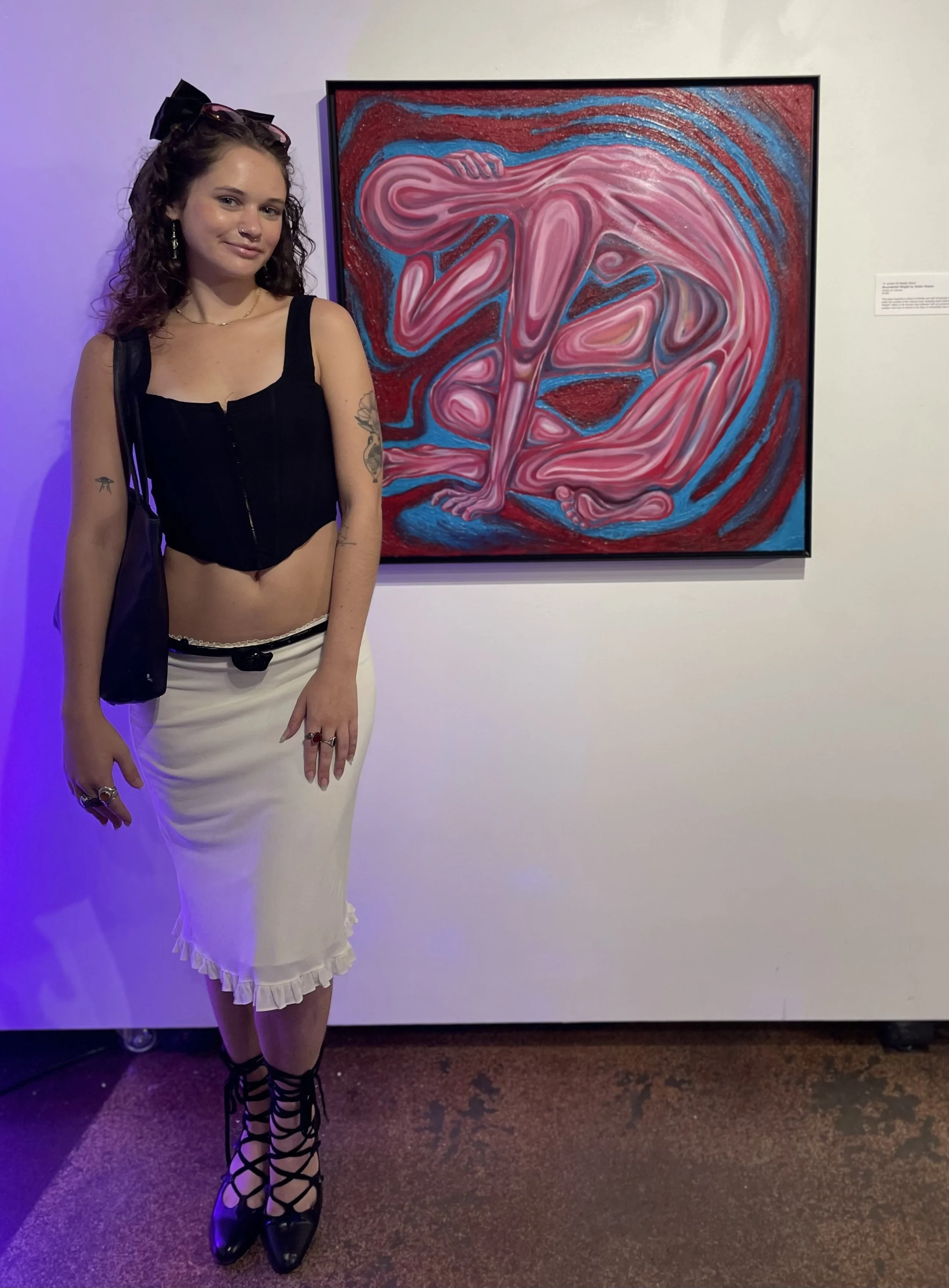 A young woman standing in front of a colorful abstract painting at an art gallery. She has curly dark hair, wears a black sleeveless top, a white skirt with a ruffled hem, and black lace-up high heel shoes.