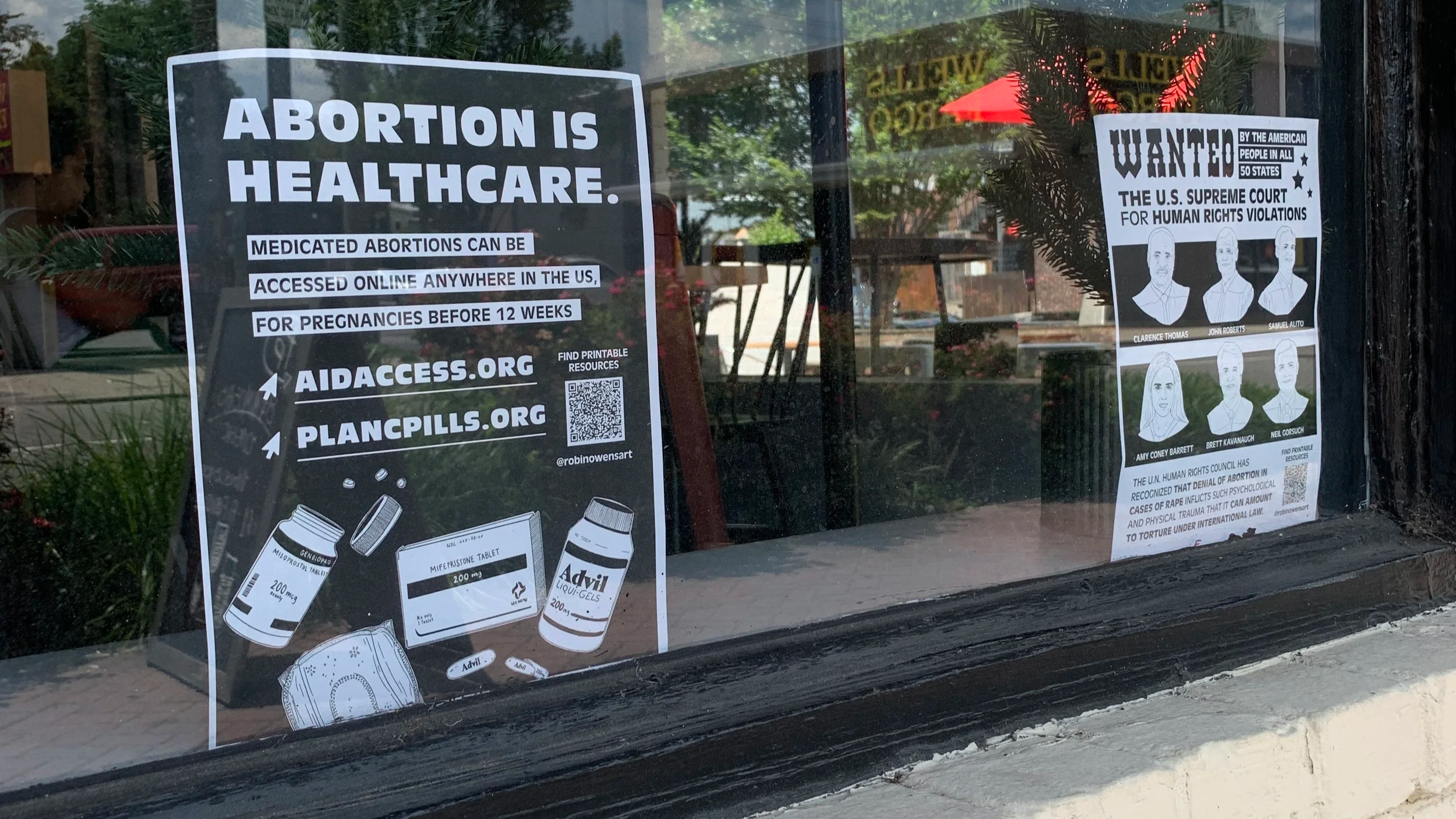 Posters on a window advocating for abortion rights and highlighting issues with the U.S. Supreme Court, including a wanted poster of Supreme Court justices and information about abortion access and human rights violations.