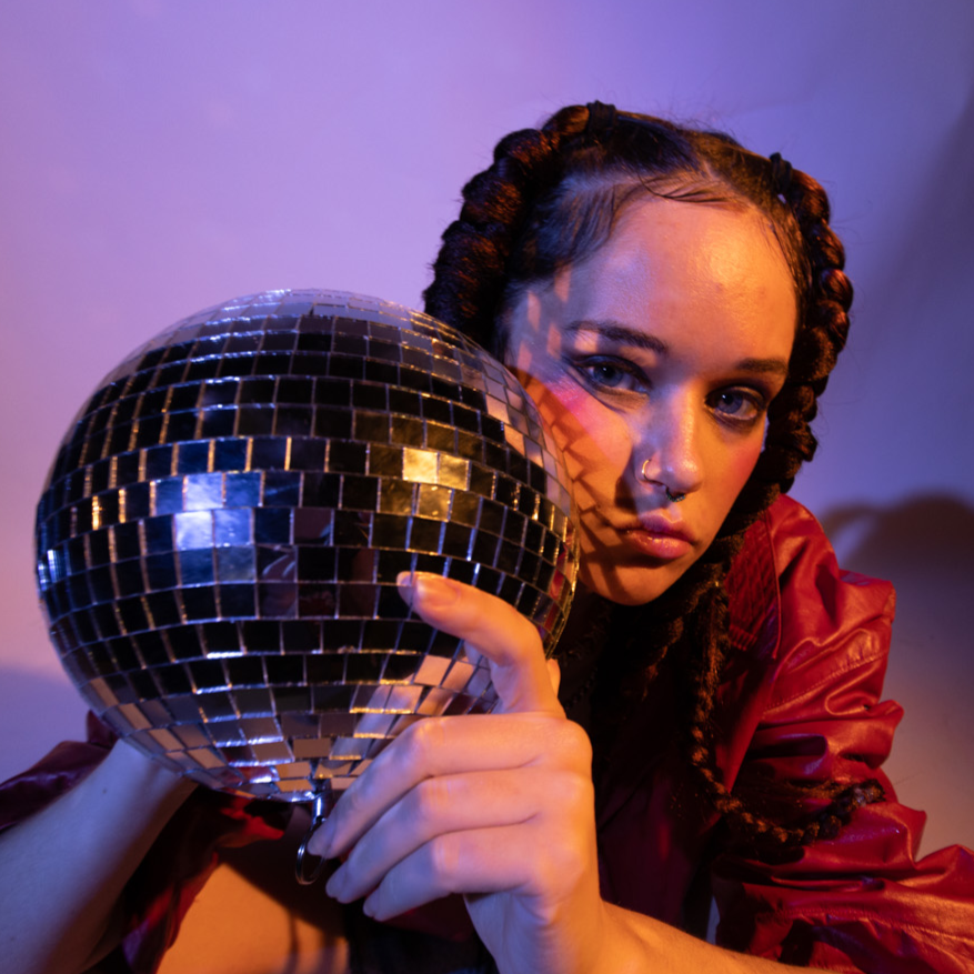 Woman with braided hair holding a disco ball in a colorful, artistic setting.