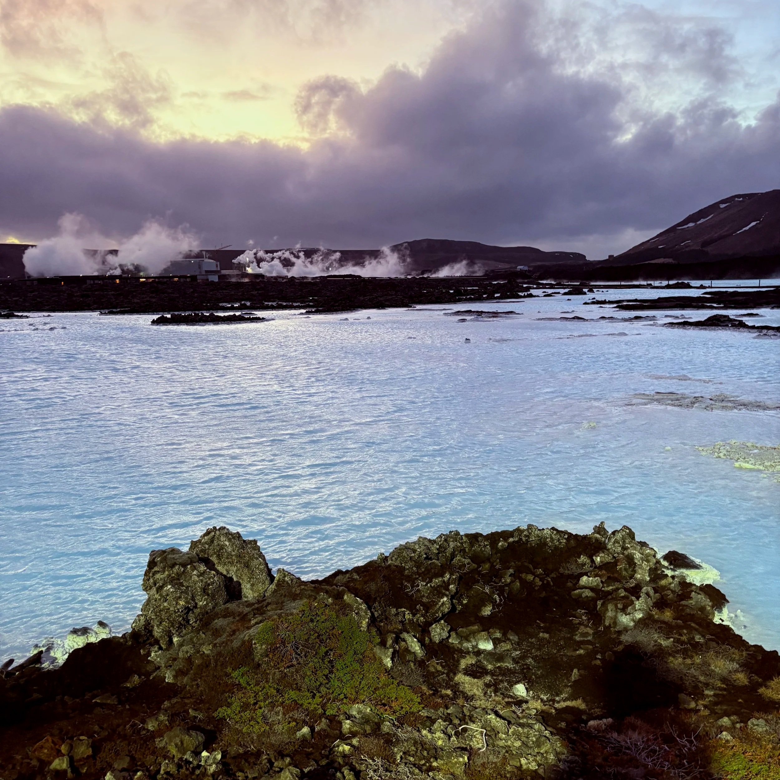 Steam rising from geothermal vents in a volcanic landscape with a body of water, rocky foreground, mountains, and cloudy sky in the background.