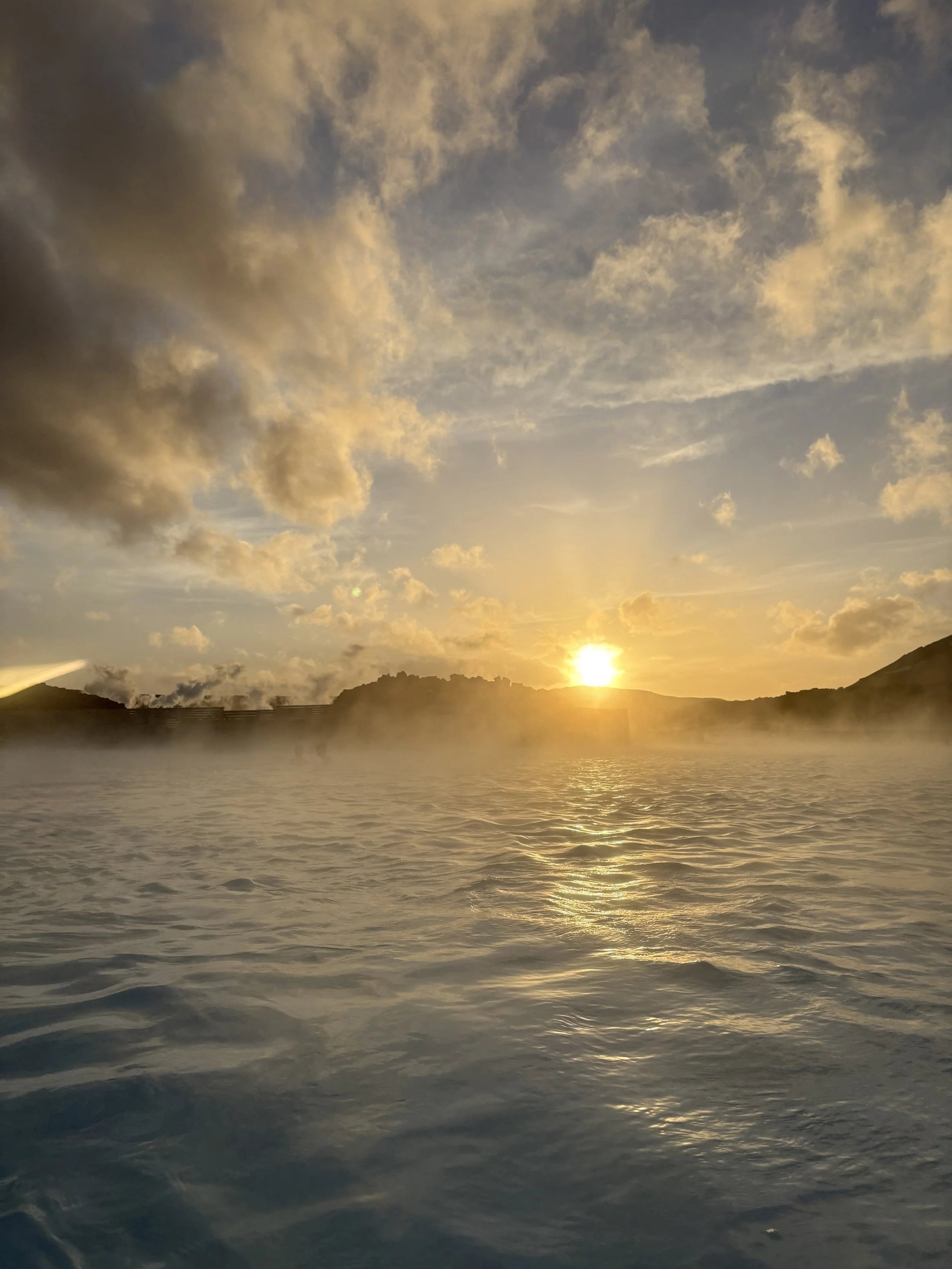 Sunrise over a body of water with mist, hills in the background, and a partly cloudy sky.