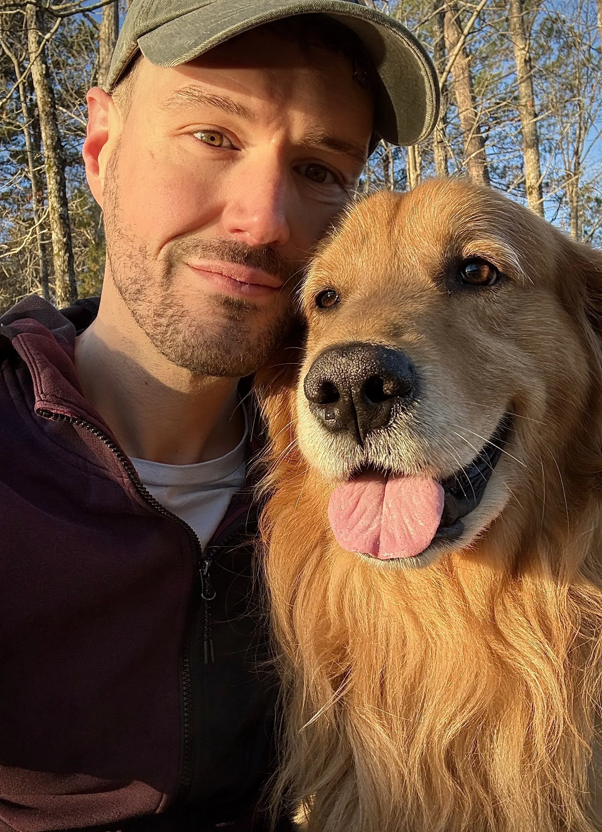 A man wearing a cap and dark jacket taking a selfie with a golden retriever dog outdoors, both appearing happy with the dog sticking its tongue out. The background shows trees and a clear sky.