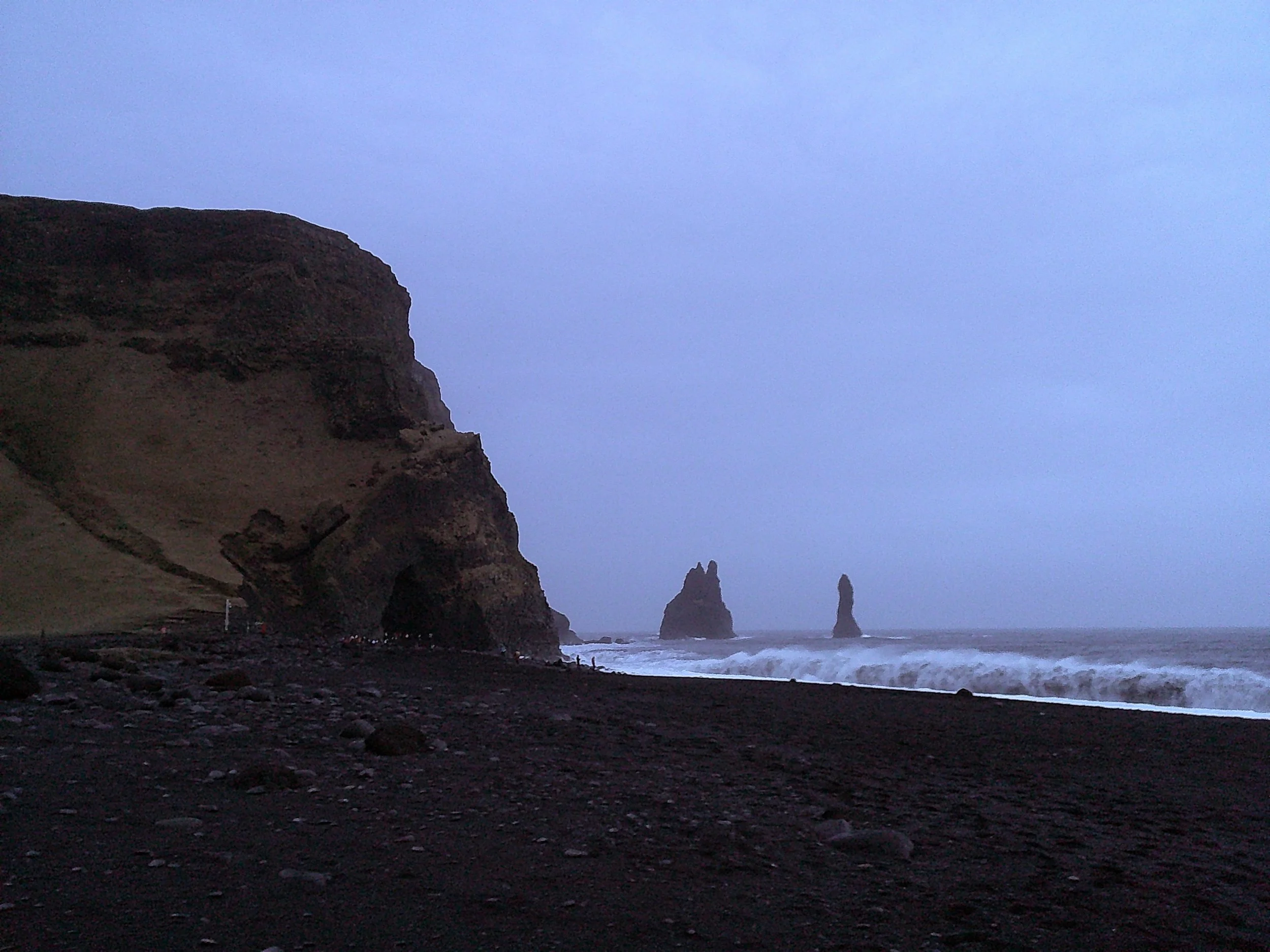 A dark sandy beach with large rock formations near the shore and an ocean with waves, a cliff on the left, and a cloudy sky.