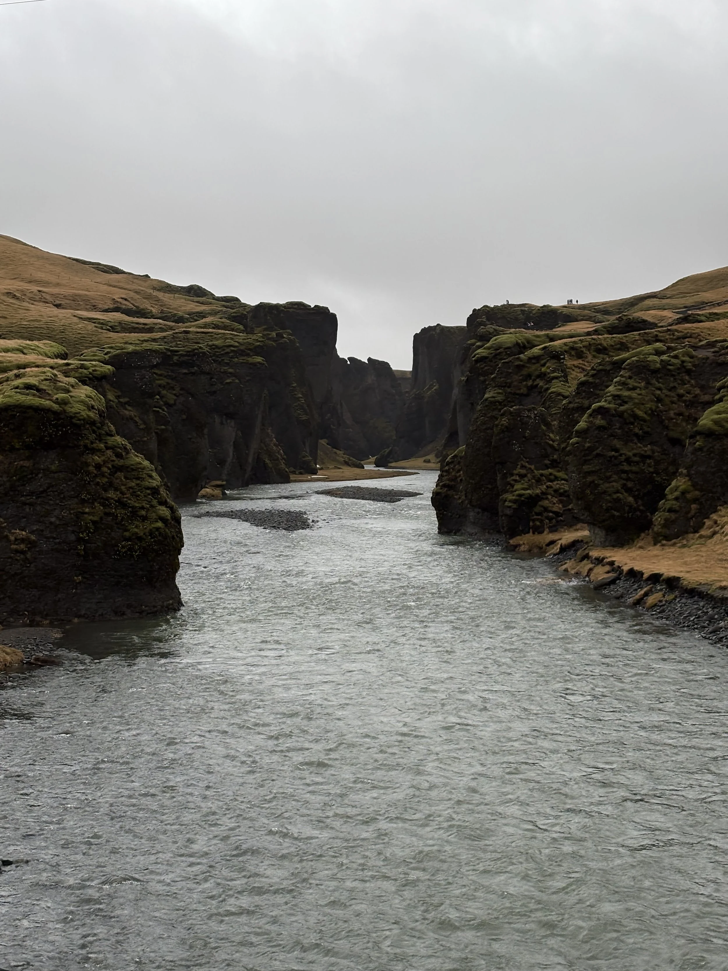 A river flowing through a canyon with moss-covered cliffs on both sides under an overcast sky.