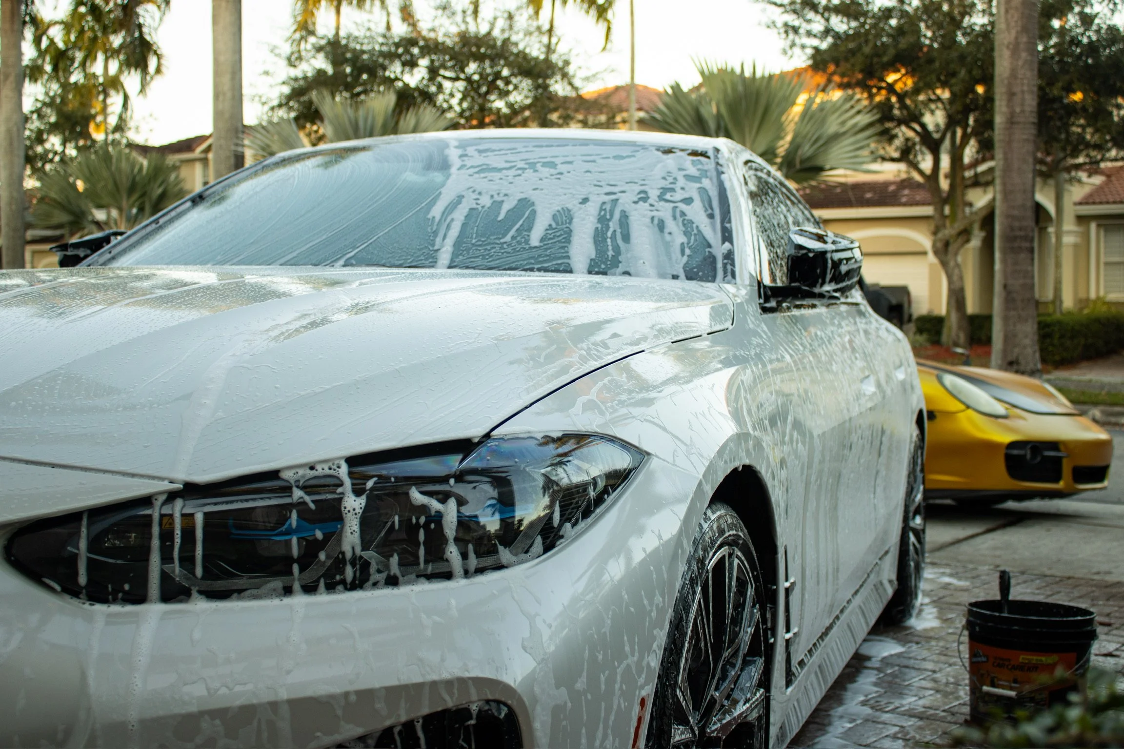 A white car being washed, covered in soap suds, with a yellow car parked in the background in a residential neighborhood with trees and houses.