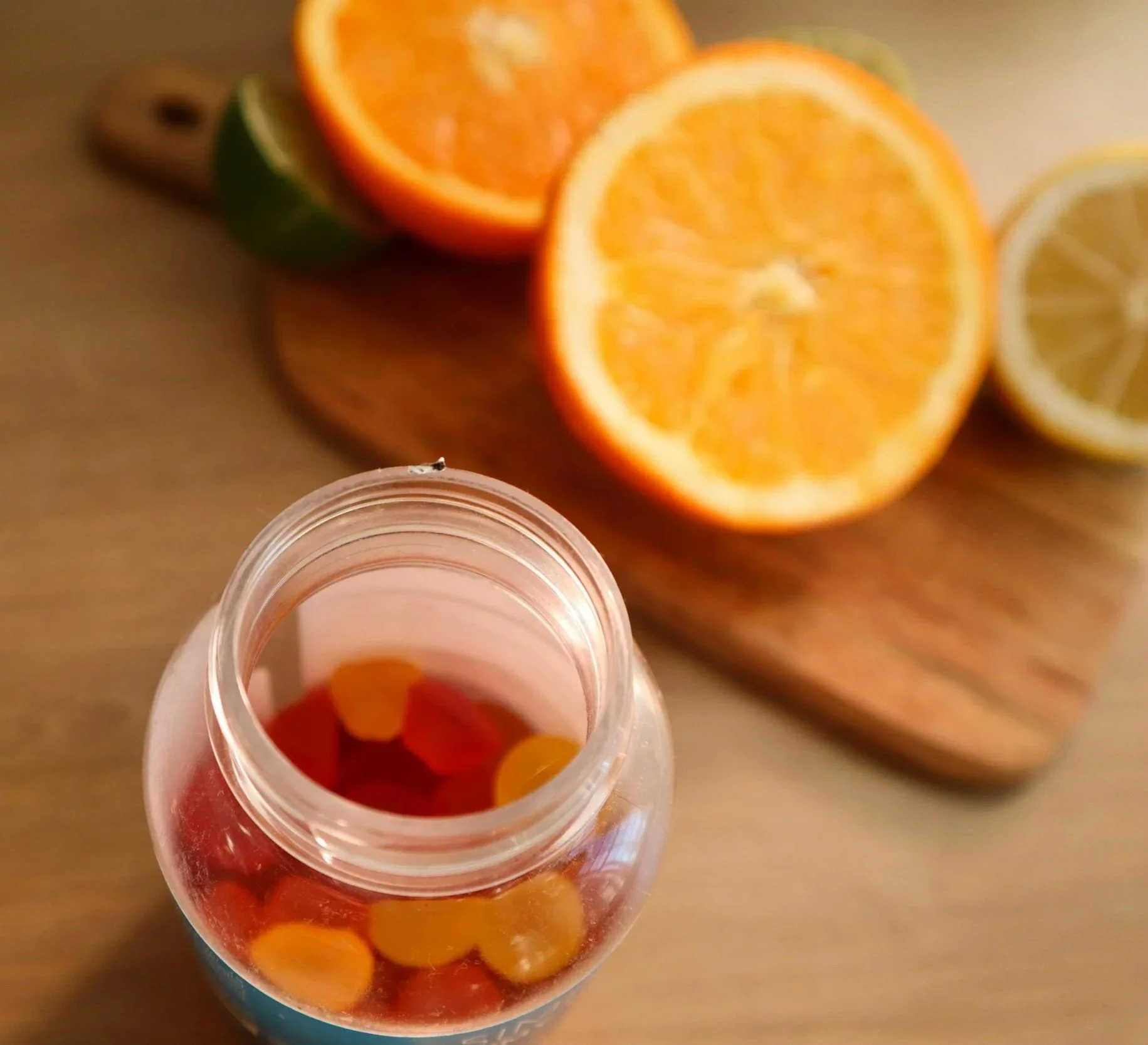 Open vitamin bottle with multi-colored medication vitamins inside, placed on a wooden surface with cut oranges, limes, and lemons in the background.