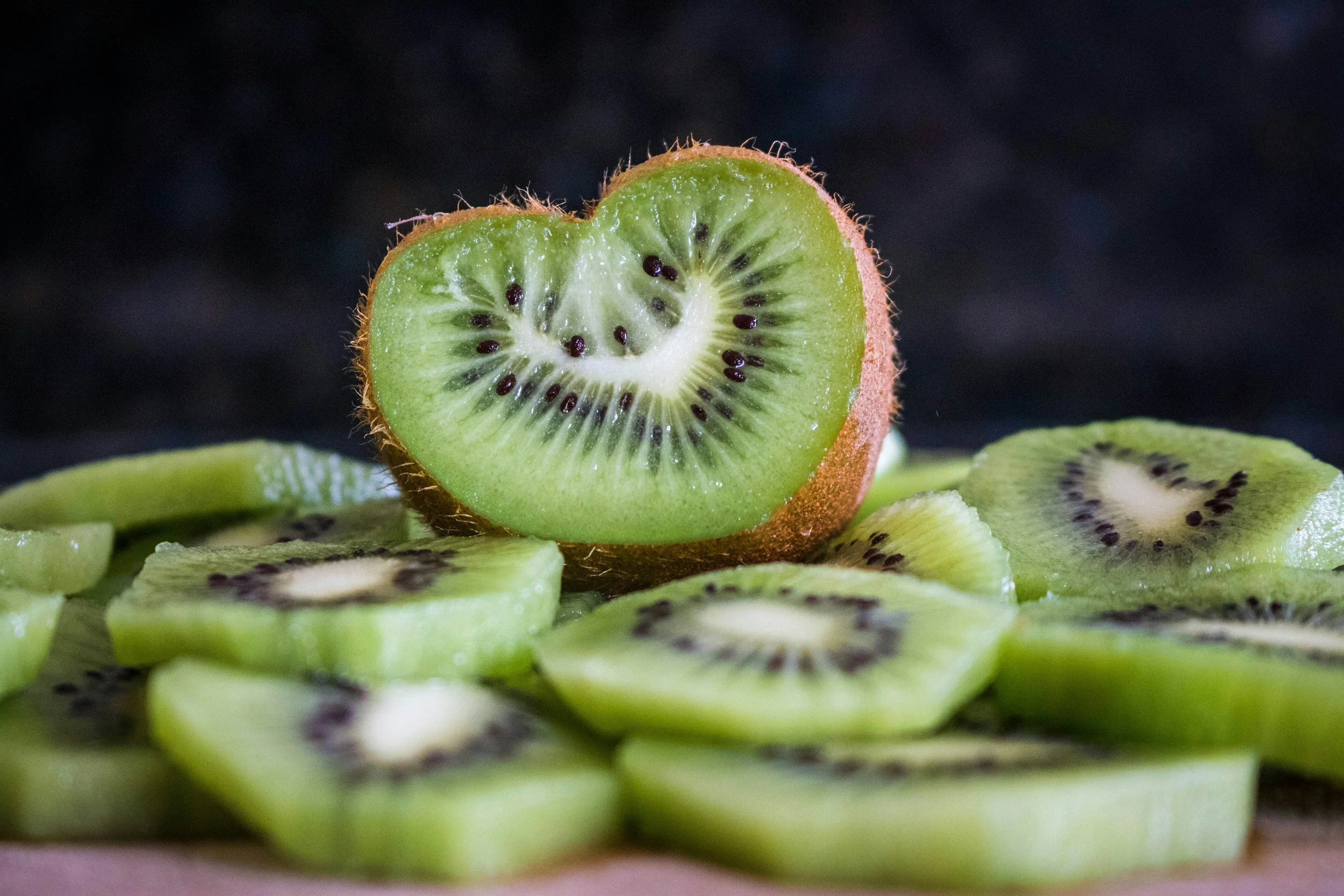 Close-up of a sliced kiwi fruit with a half kiwi on top, surrounded by more slices of kiwi on a dark background.