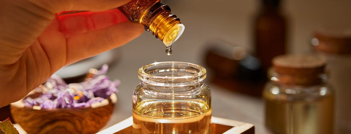 A person holding a small amber-colored bottle, pouring liquid into a glass jar of golden liquid, with purple flowers in the background.