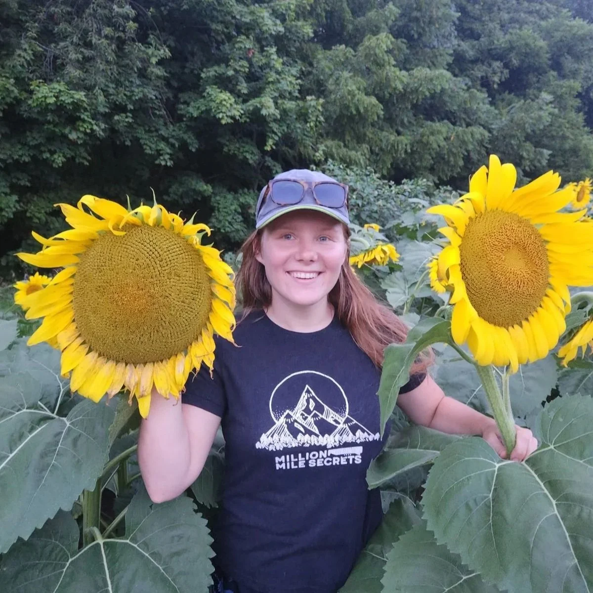 A young woman with long red hair wearing sunglasses on her cap, a black Mountain of the Moon T-shirt, and holding two large blooming sunflowers, standing in a sunflower field with trees in the background.